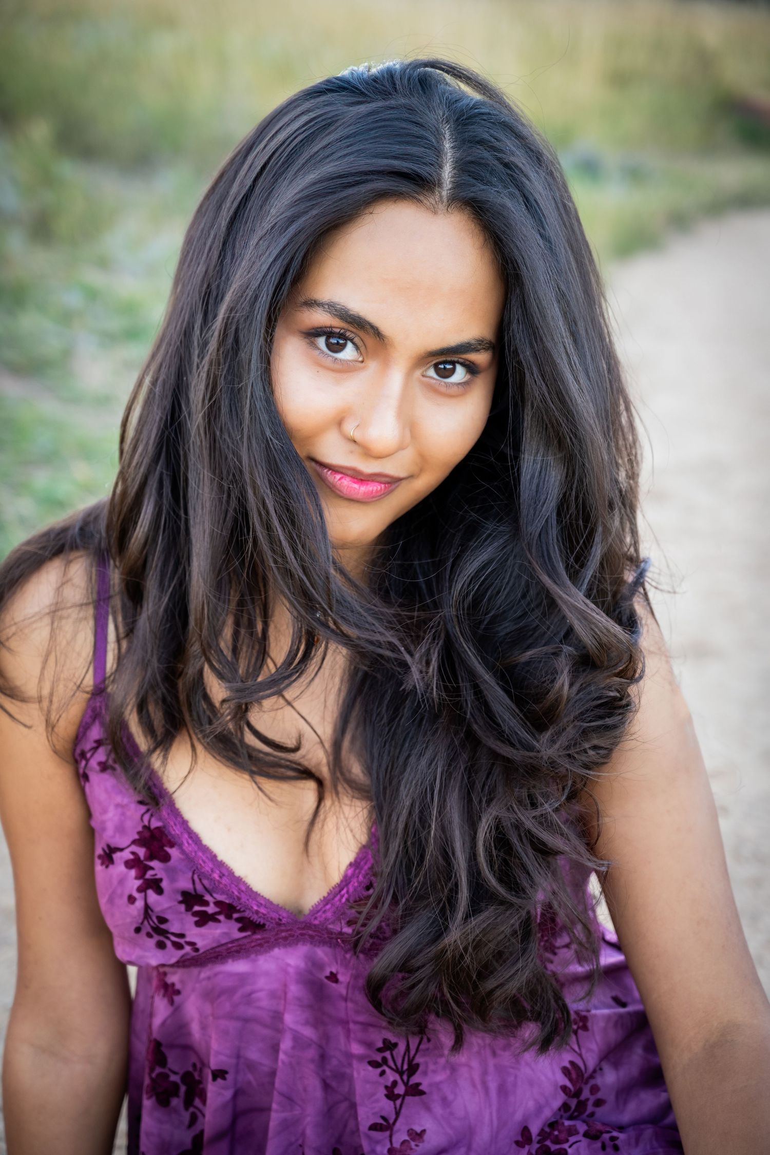 Young woman with long dark wavy hair wearing a purple floral dress outdoors on a dirt path.