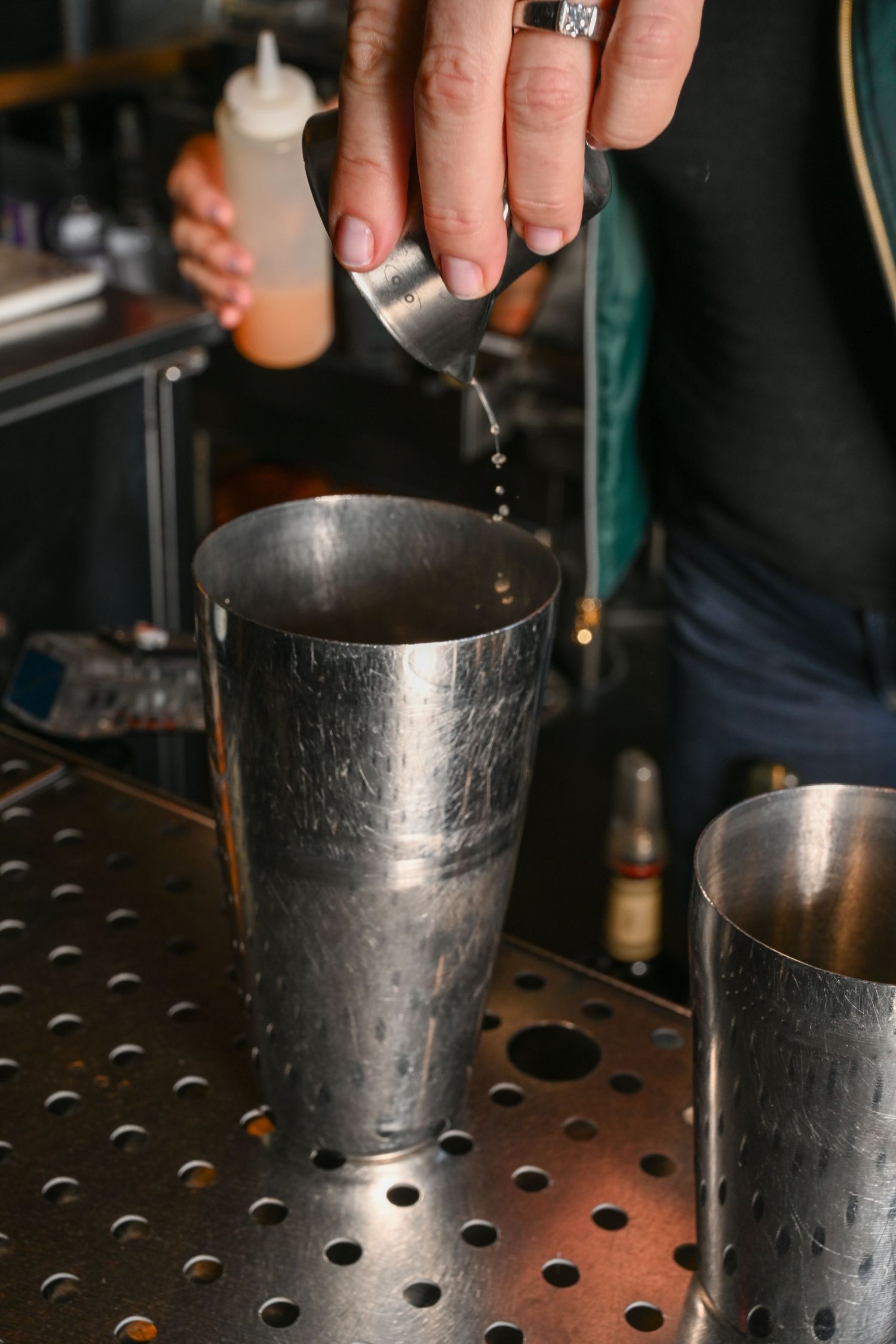 Bartender pouring a measured shot into a metal cocktail shaker on a perforated bar mat.
