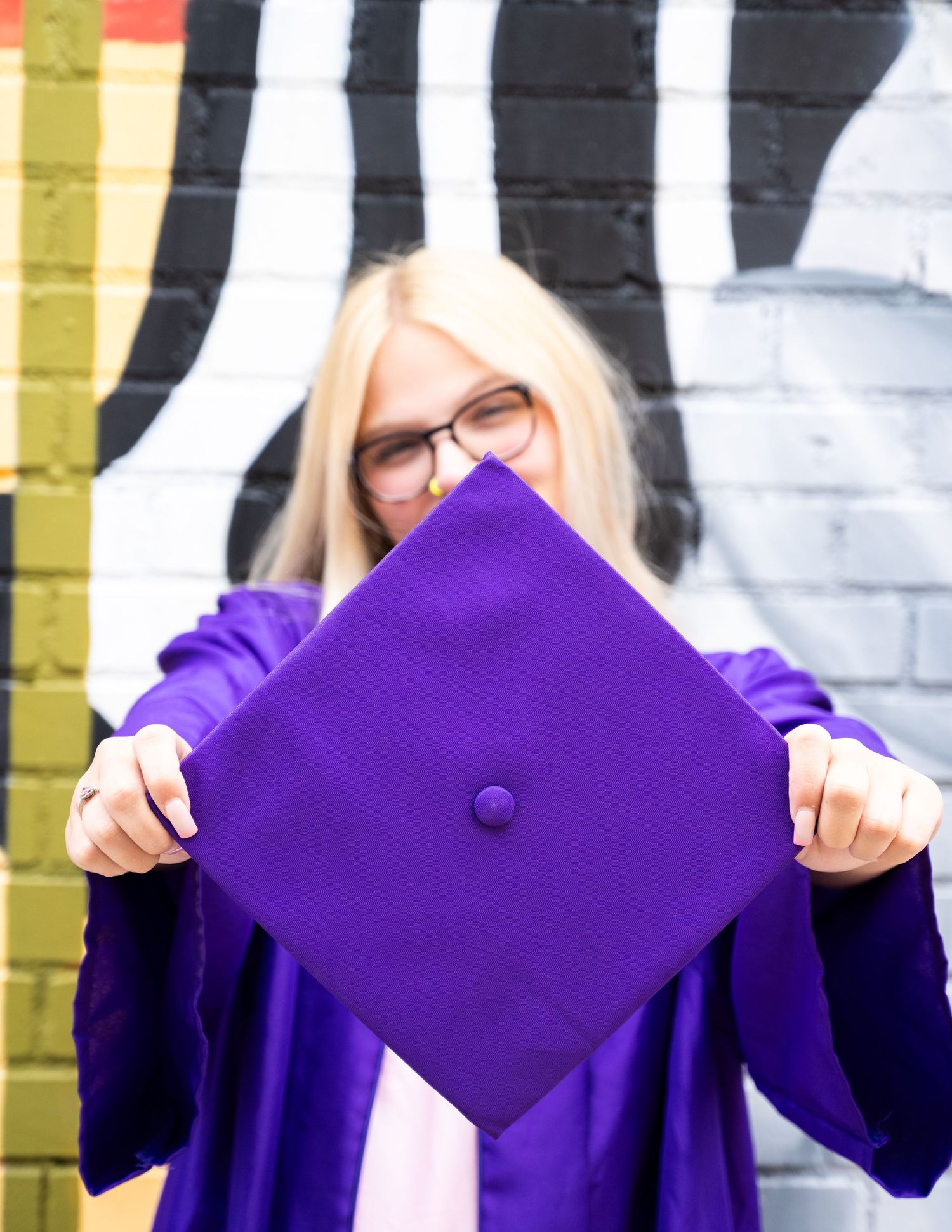 Blonde graduate in purple cap and gown holding mortarboard in front of graffiti wall.