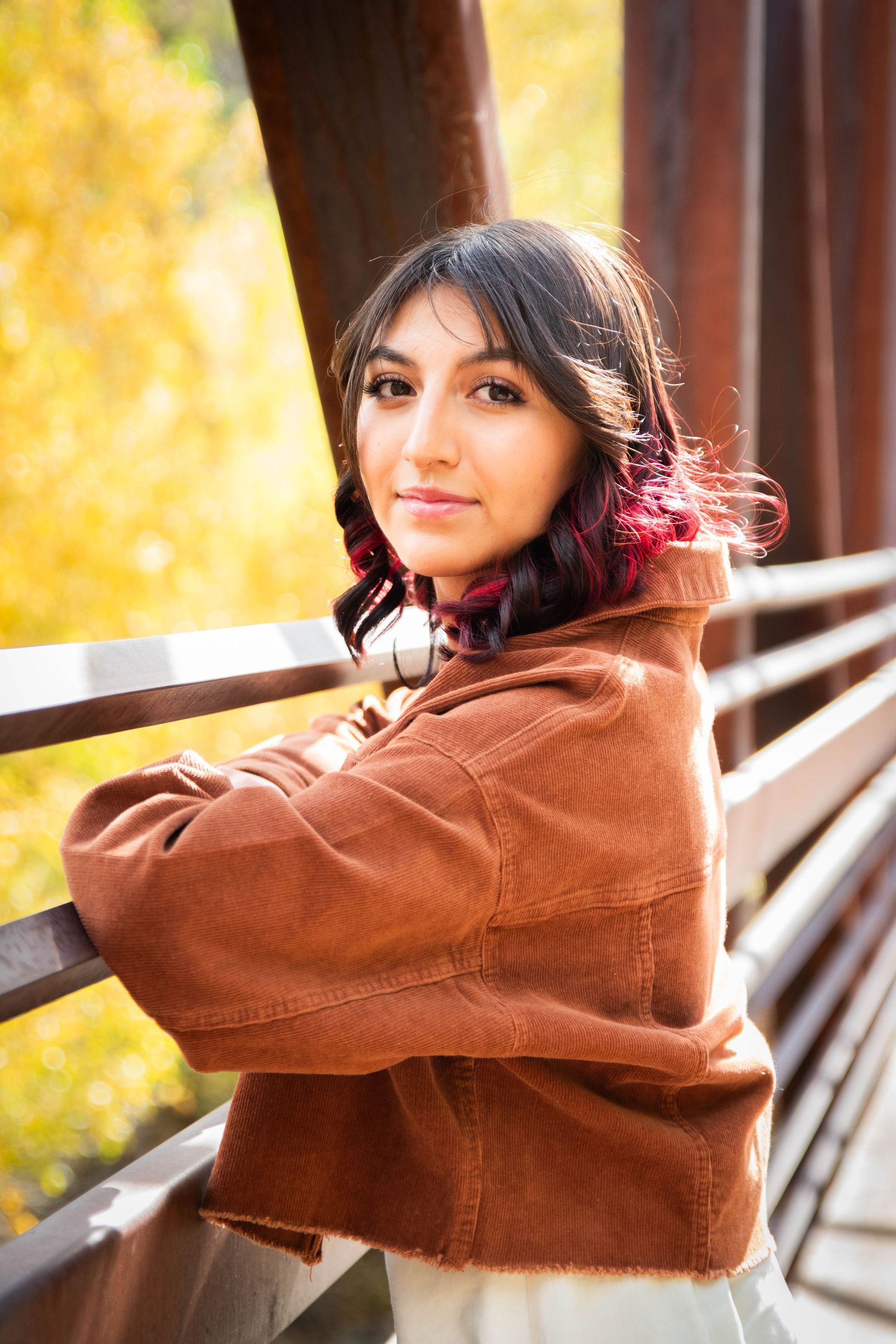 Young woman with pink-highlighted hair in brown jacket leans on bridge railing in autumn setting.