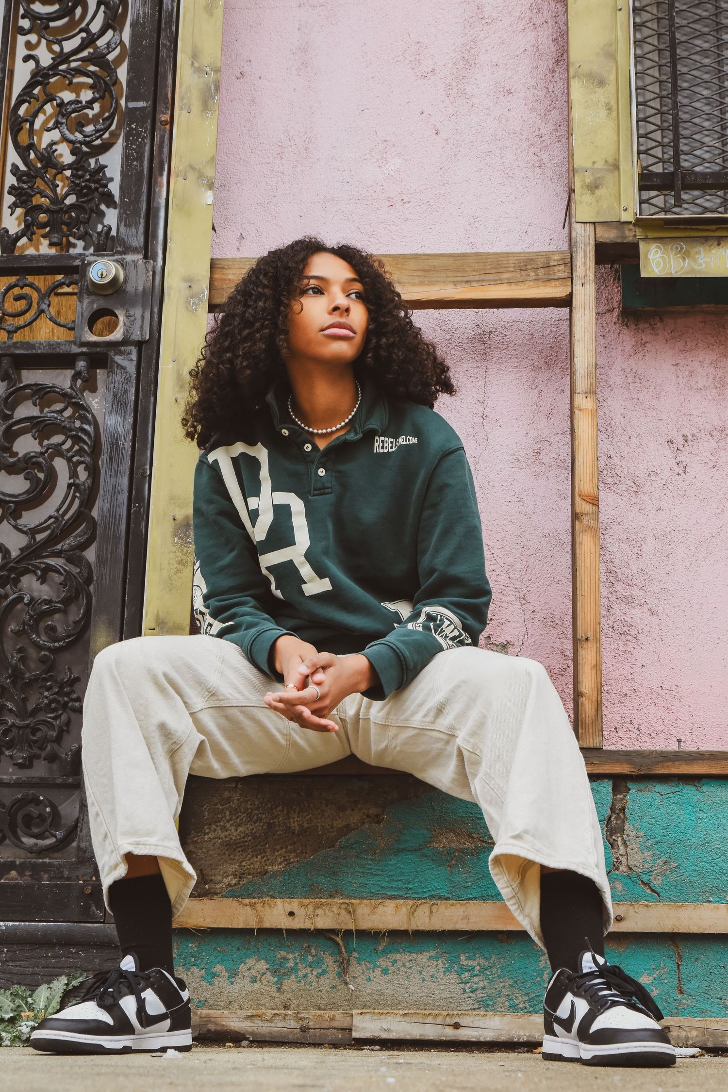 Young woman with curly hair in green sweatshirt sits on steps before colorful urban building.