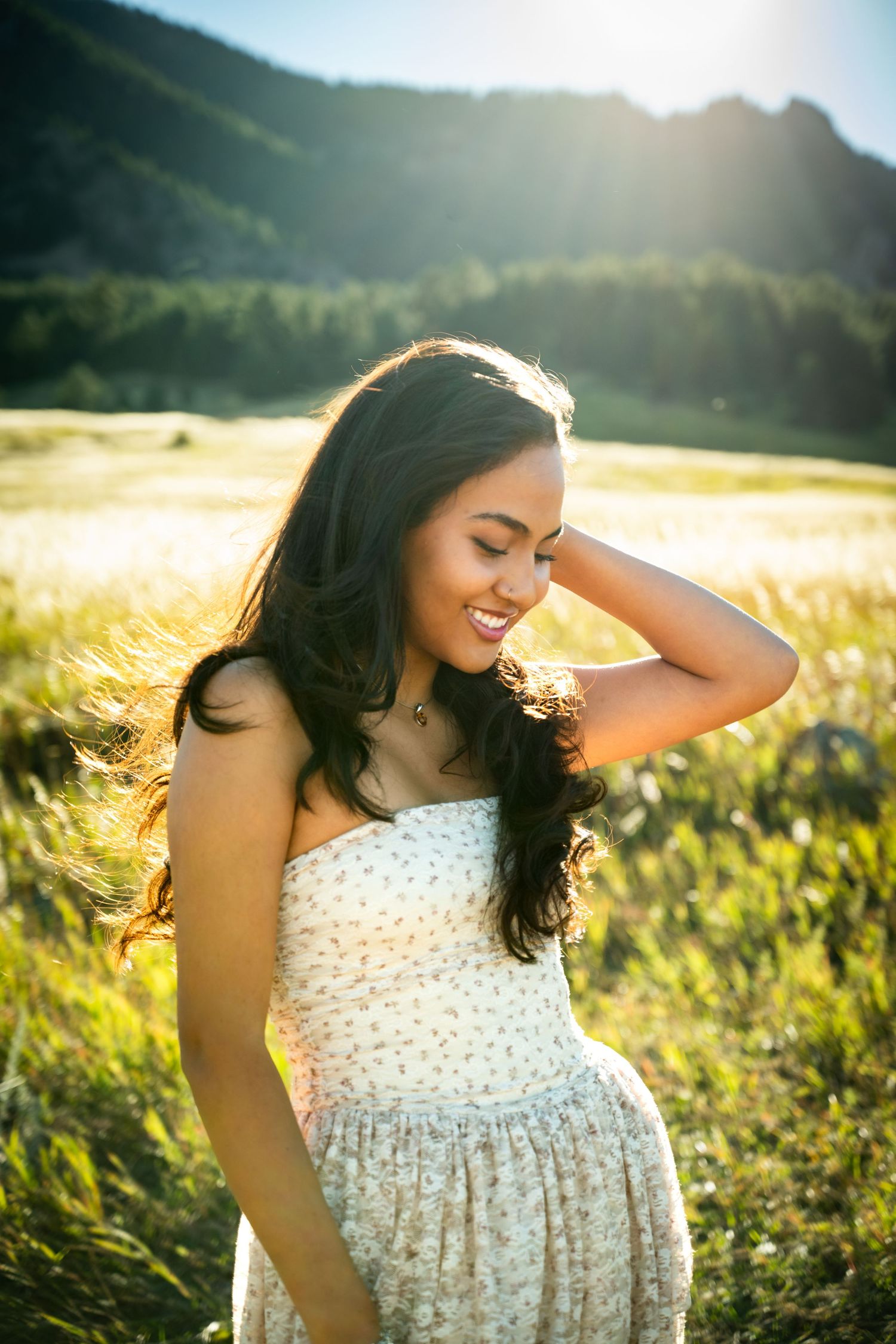 Young woman with long dark hair smiling in golden sunlit meadow wearing white floral dress.