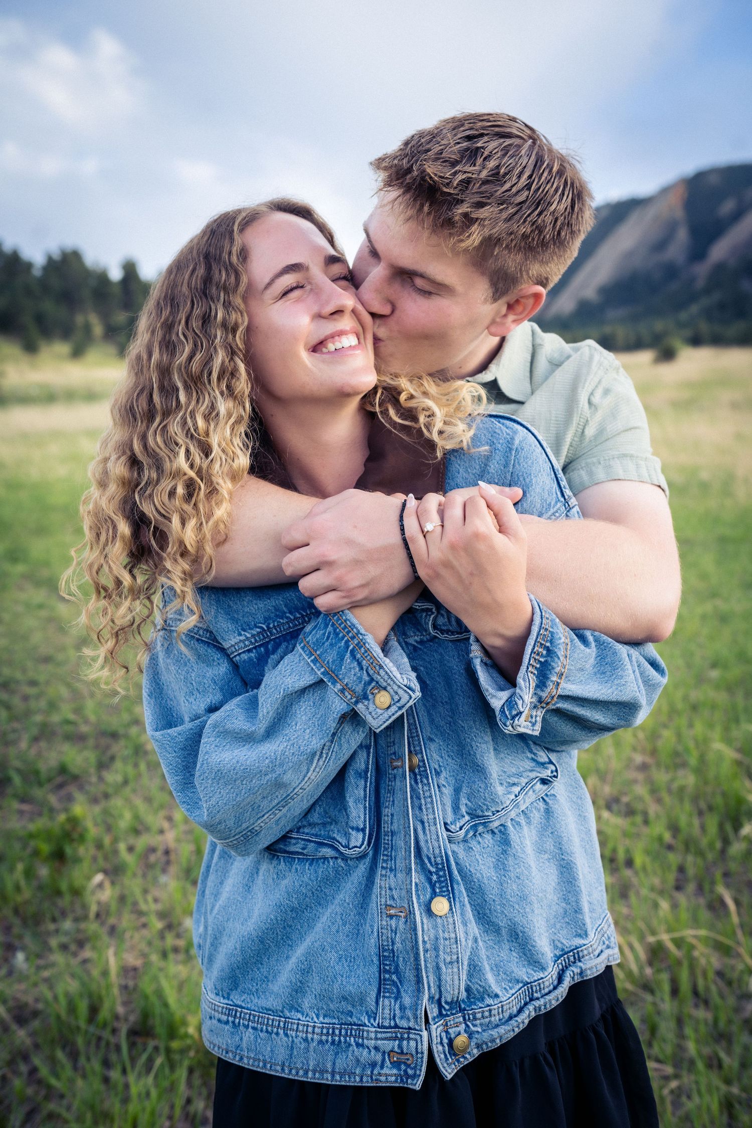 Young man embracing woman from behind, kissing her cheek, smiling in a mountain meadow.