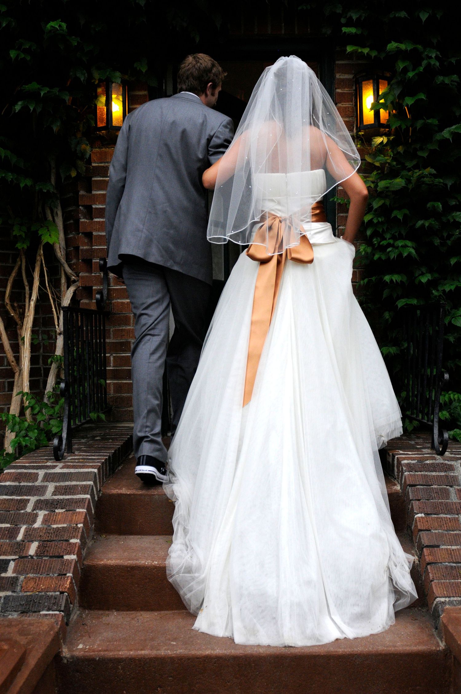 Bride and groom ascending brick steps, bride wearing white gown with orange sash and veil.