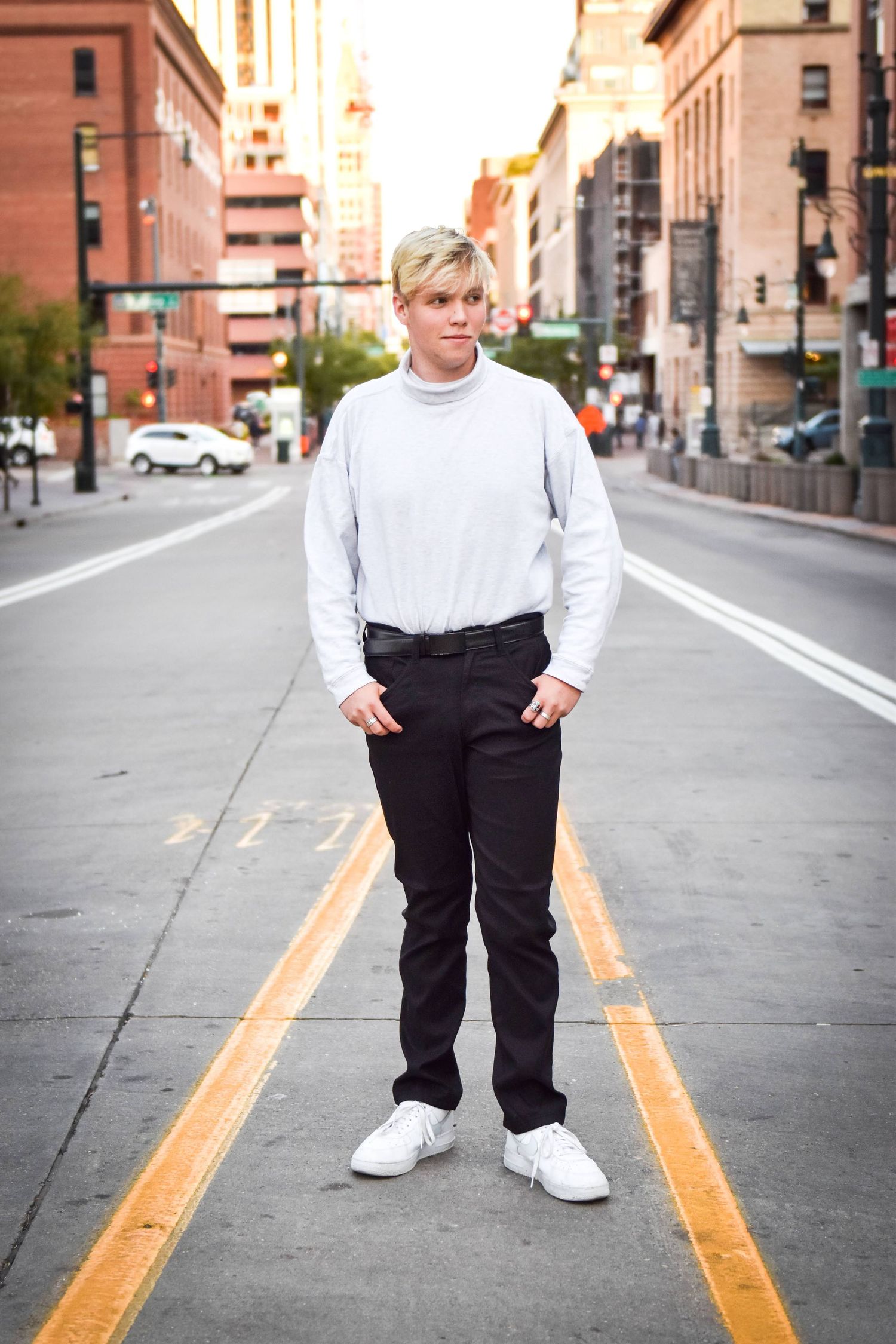 Blonde teen in white turtleneck and black pants standing on an urban city street.