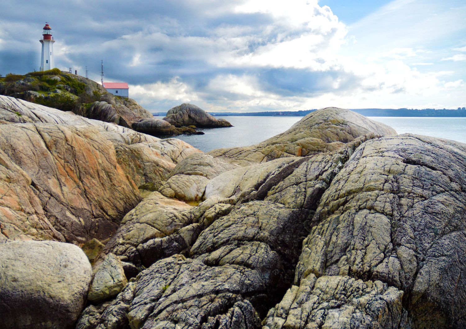 Rocky coastline with lichen-covered granite boulders leading to a white lighthouse under dramatic cloudy skies.