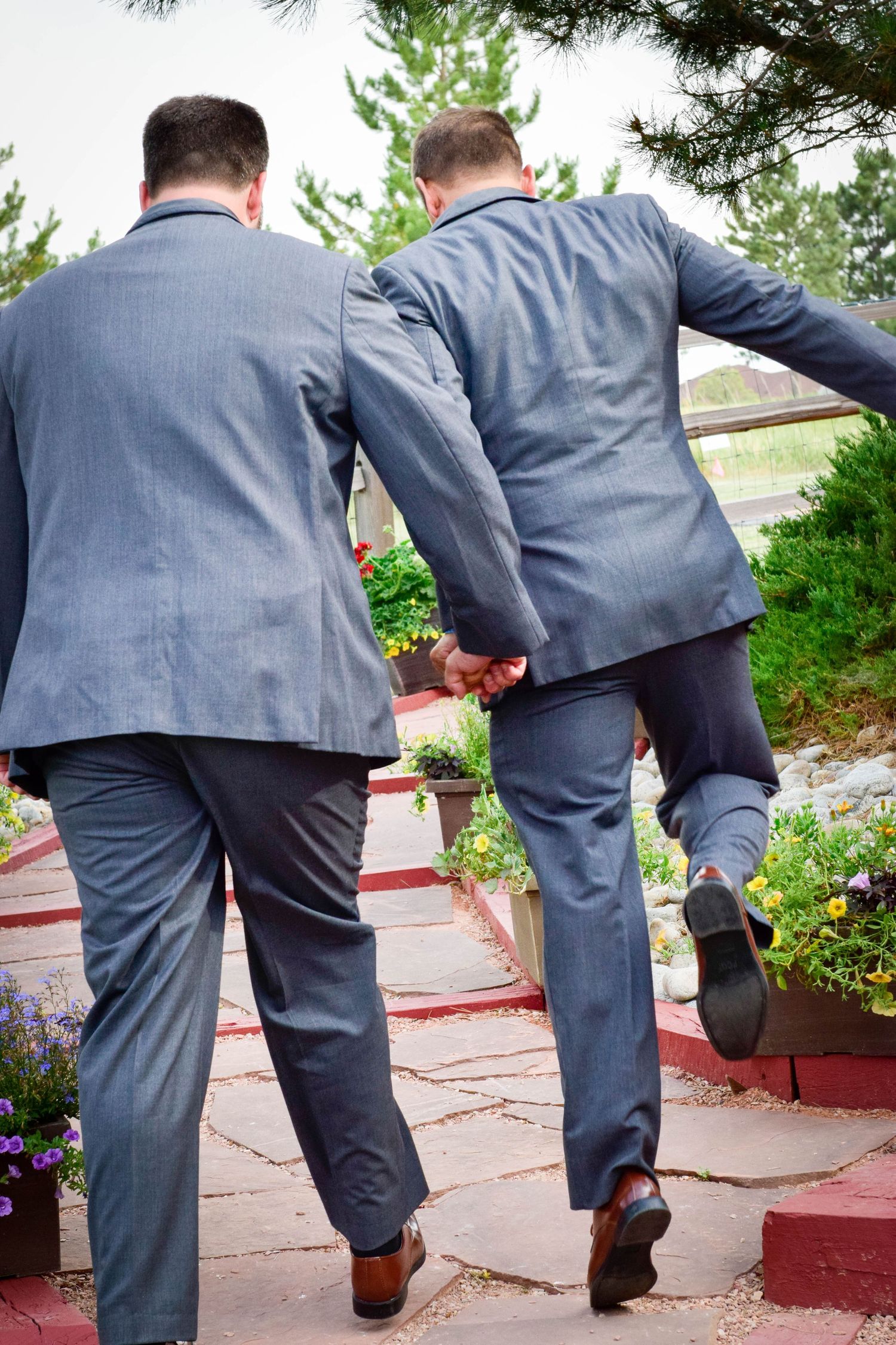 Two grooms in matching gray suits walk hand-in-hand up stone steps in a garden setting.