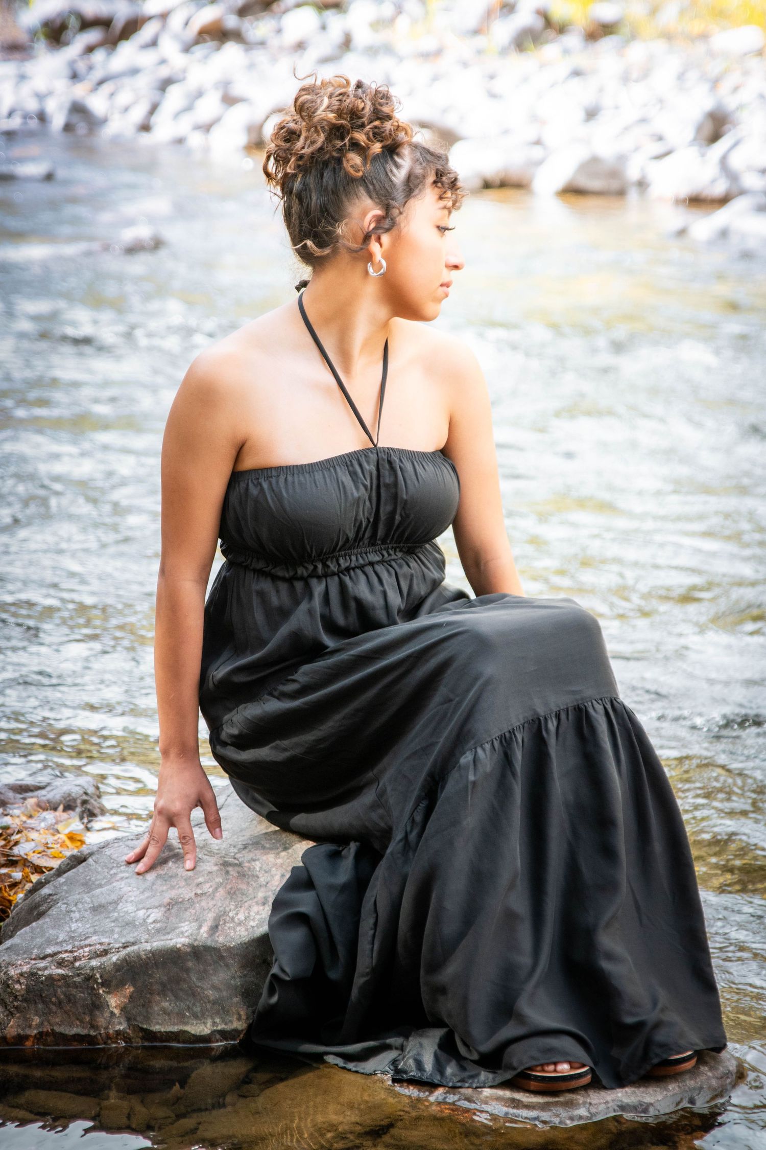 Woman in elegant black maxi dress sitting on rocks beside a flowing river.