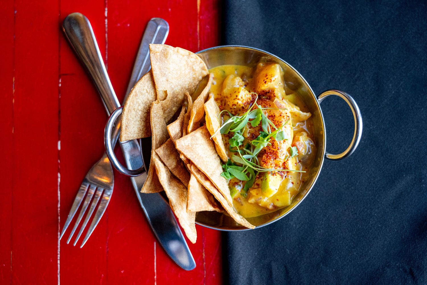 Overhead view of curry dish with flatbread chips in brass bowl on red and blue surface.
