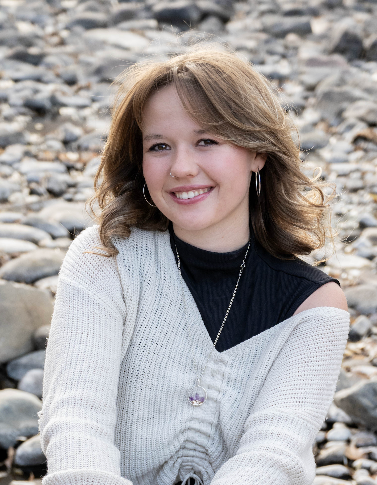 Smiling teen girl in white cardigan sitting on rocky beach with layered necklace.