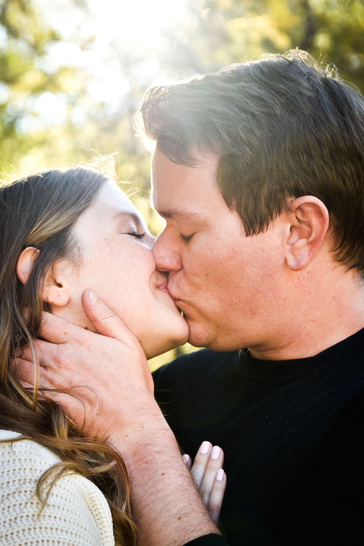 Close-up of couple sharing a romantic kiss outdoors with soft sunlight filtering through trees behind them.