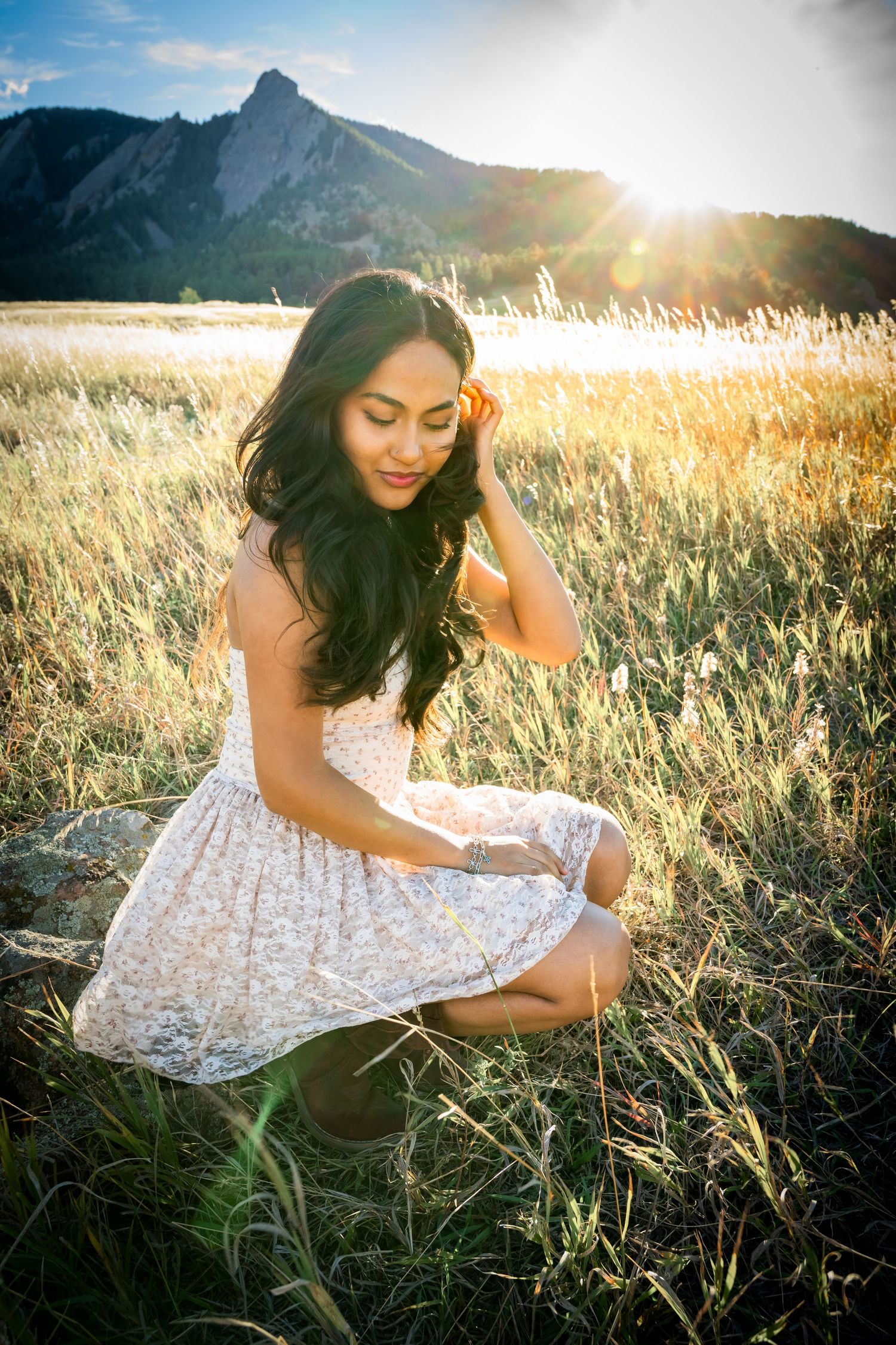 Young woman in floral dress sitting in golden meadow with mountain backdrop and sunflare.