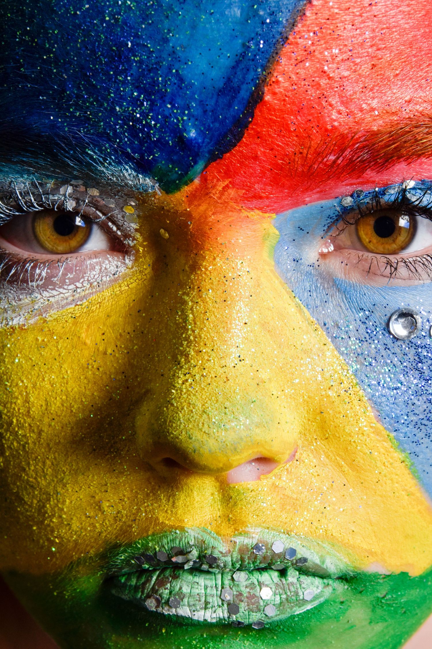 Close-up of a woman with vibrant multicolored face paint in blue, red, yellow, and green with rhinestone accents.