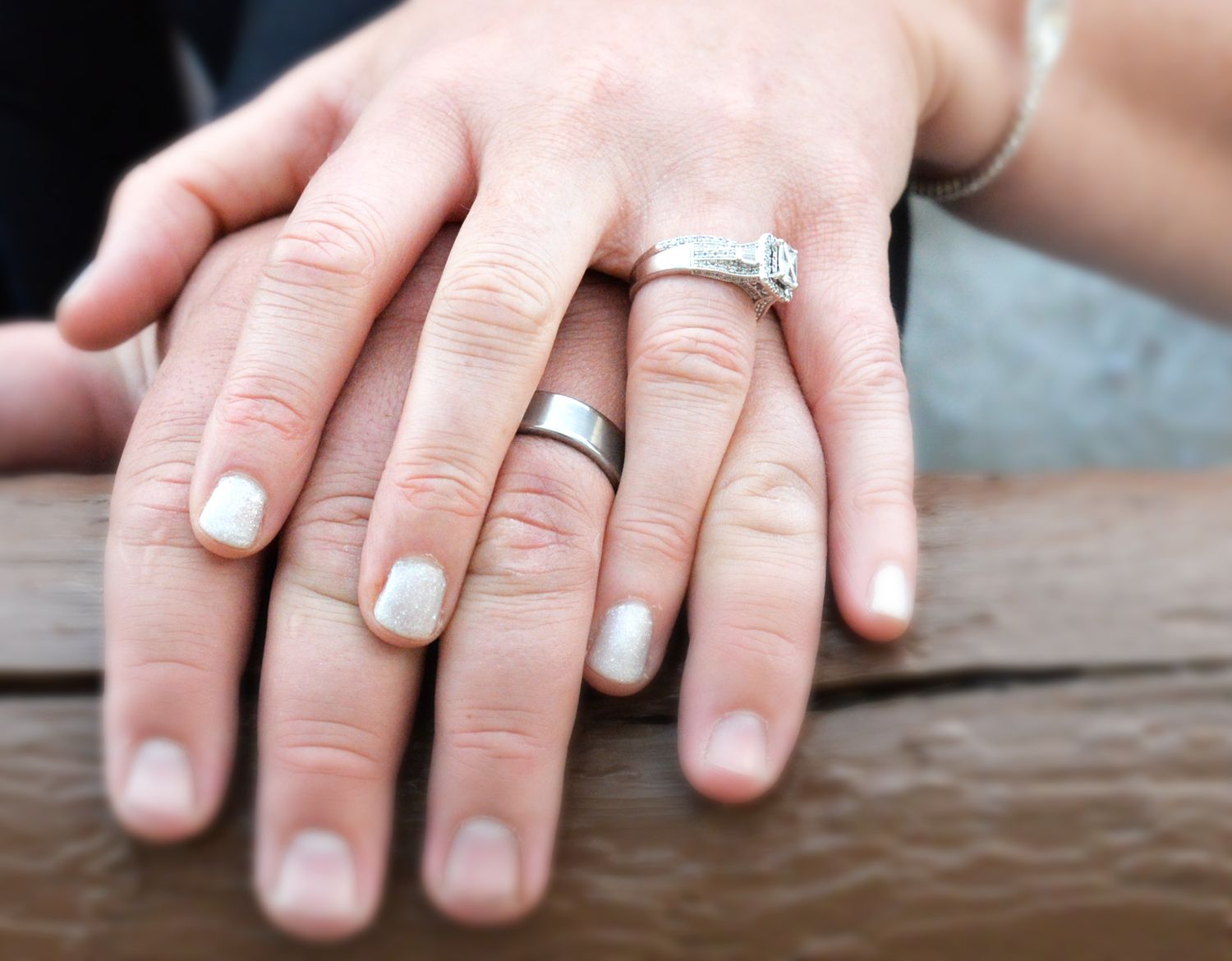 Newlywed couple displays their wedding rings, a silver band and diamond ring, resting on a wooden surface.