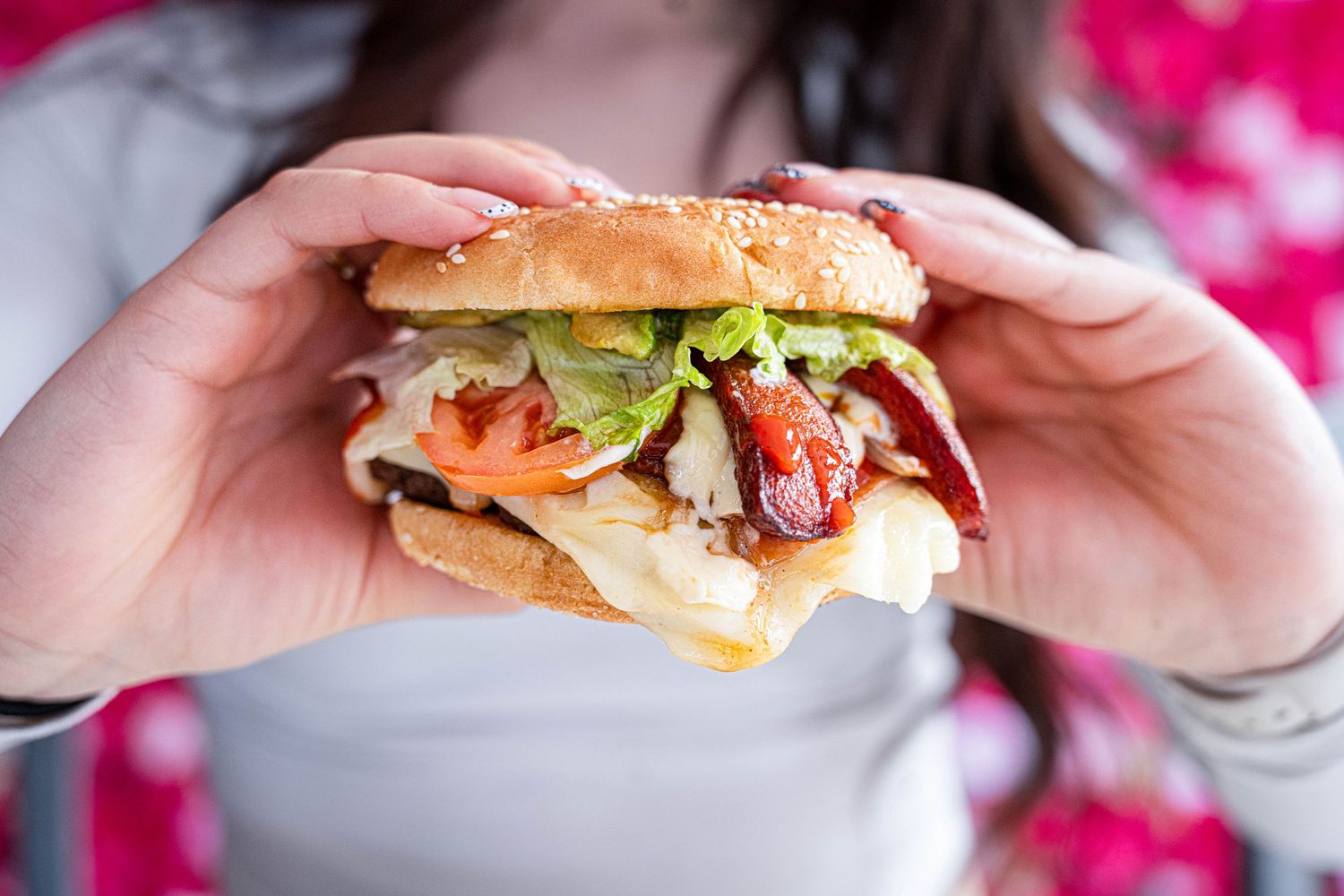 Person holding a large sandwich with lettuce, tomato, and bacon on a sesame seed bun.
