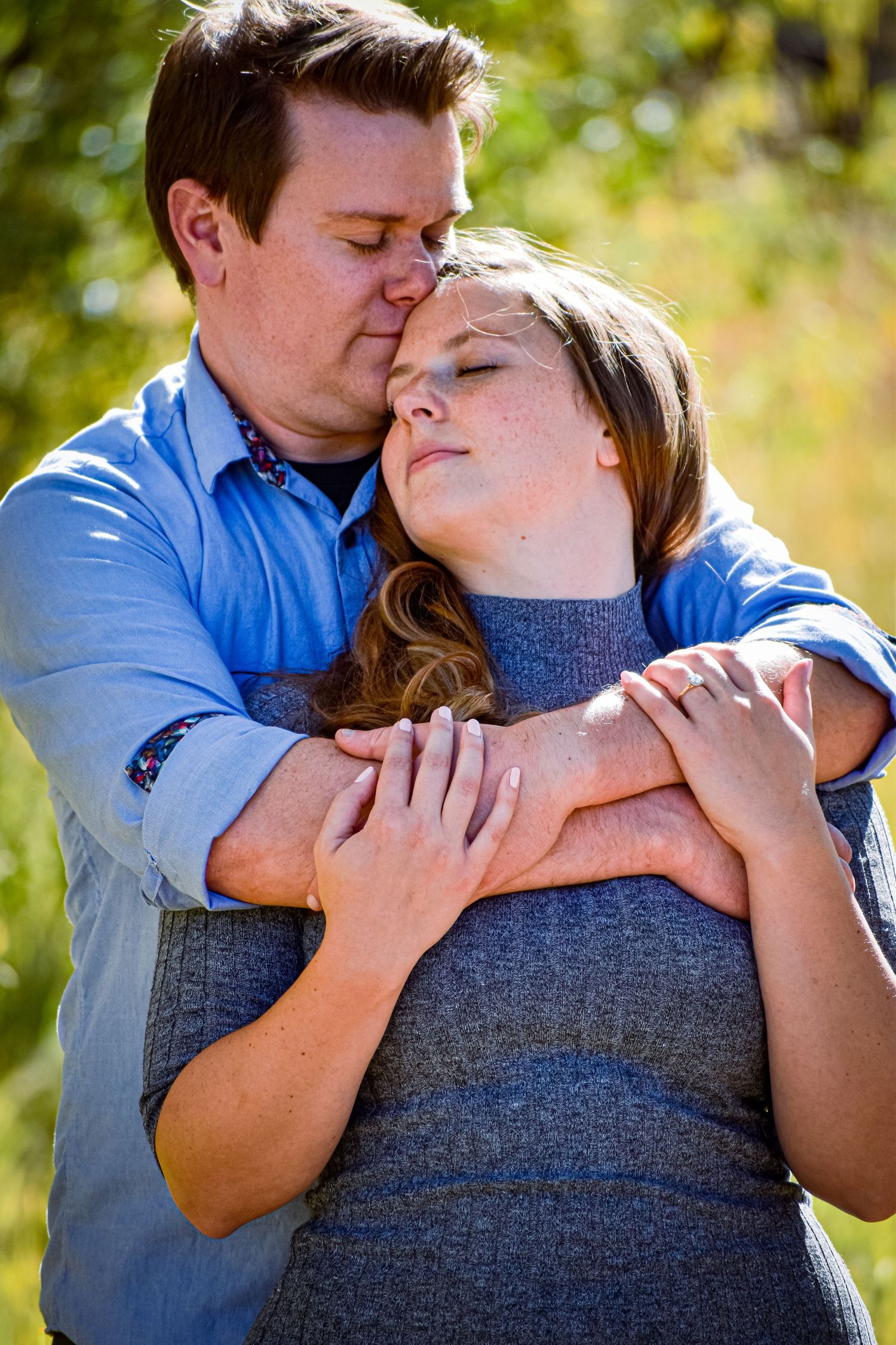 Romantic couple embracing outdoors, man in blue shirt tenderly kissing womans head in natural setting.