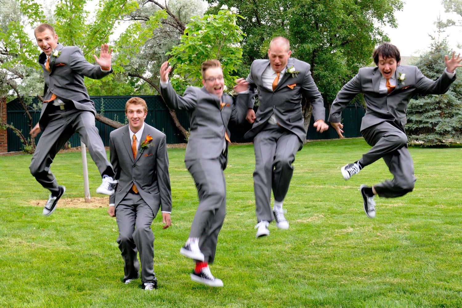 Groomsmen in gray suits jumping joyfully on green grass during a wedding celebration outdoors.