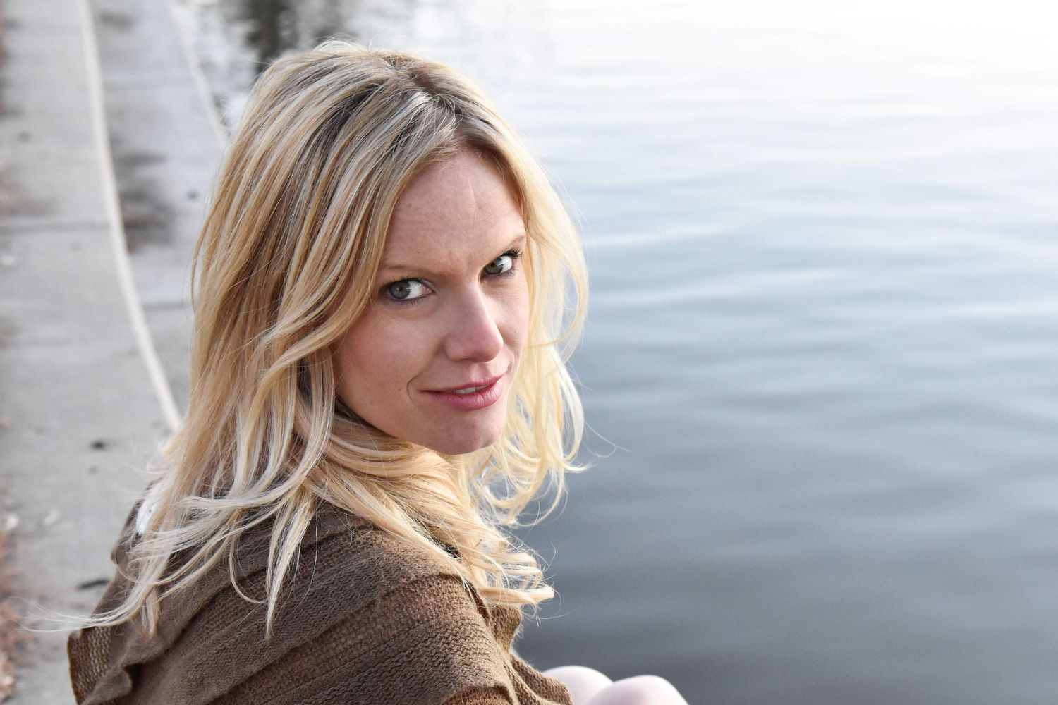 Blonde woman in brown sweater looking back over shoulder near calm waterfront.