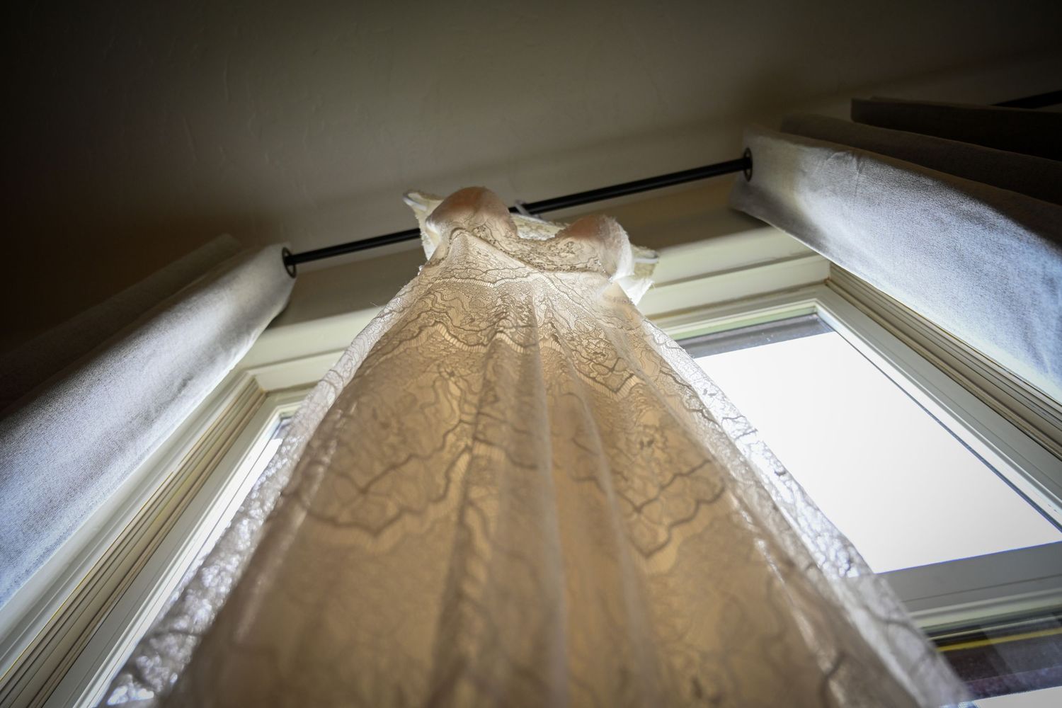 Elegant lace wedding dress hanging in a sunlit window, viewed from below.