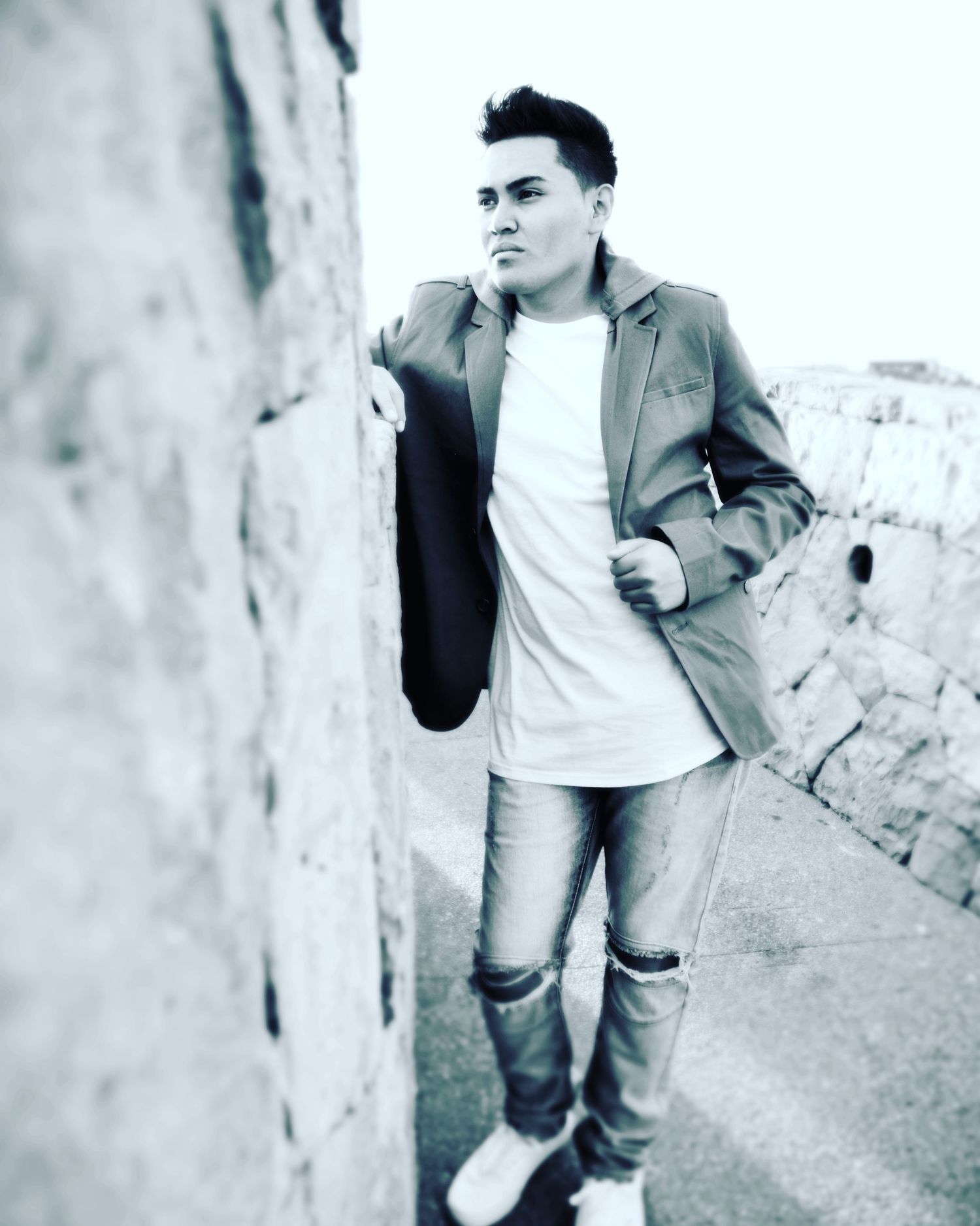 Young man in blazer and white shirt leans against stone wall, posing confidently in a stylish black and white photo.
