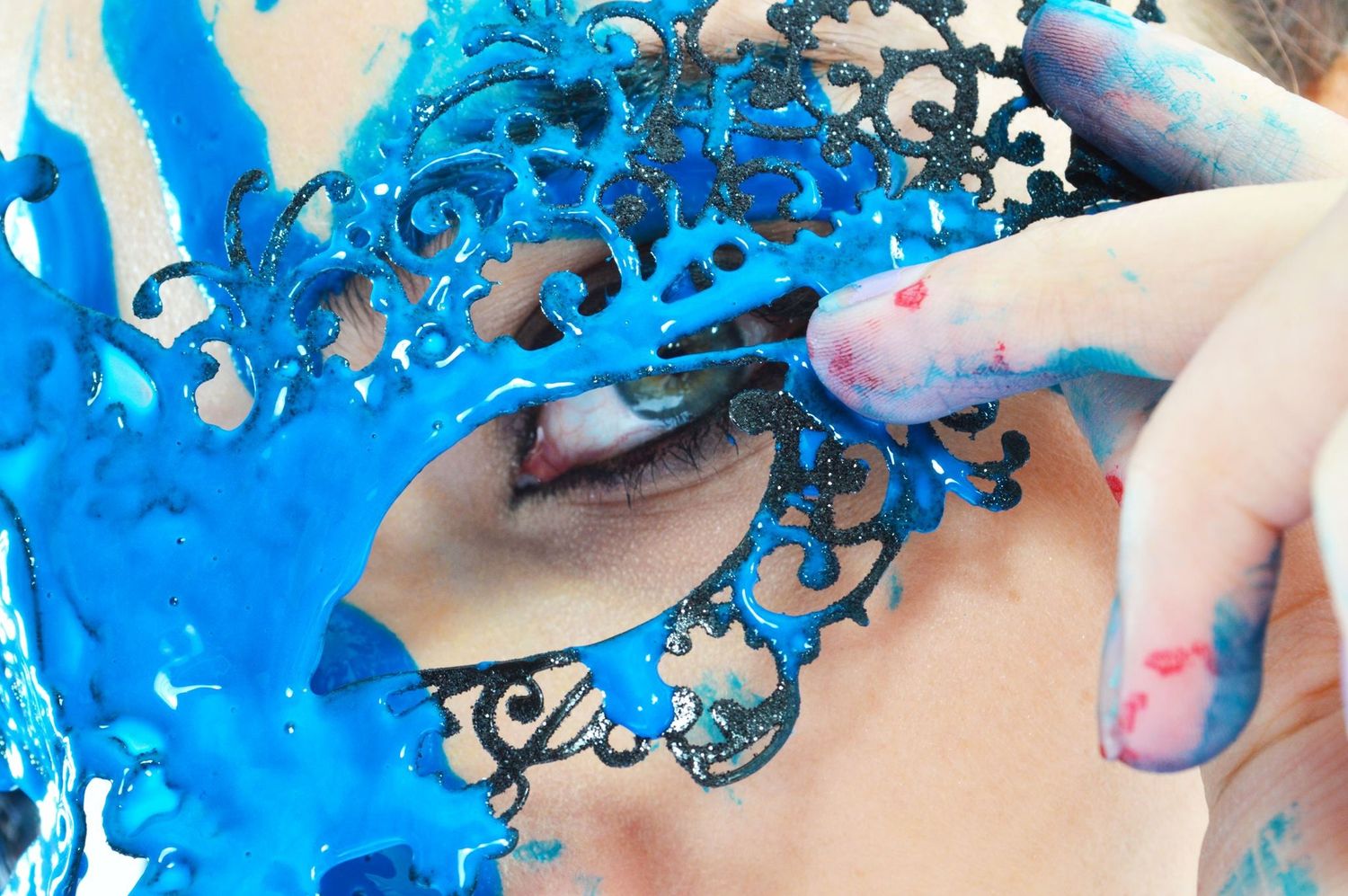 Woman wearing an ornate blue lace masquerade mask with paint-covered fingers touching her face.