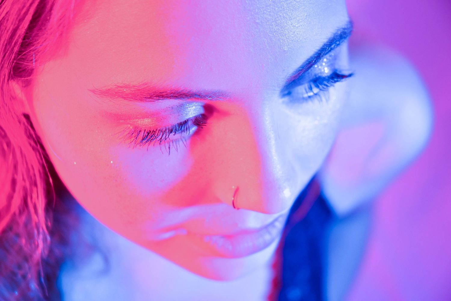 Close-up portrait of a young woman with a nose ring under dramatic pink and blue neon lighting.