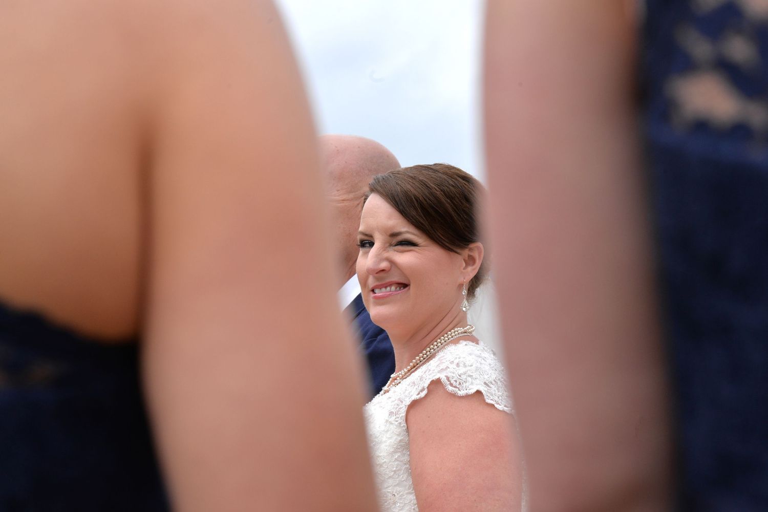 Smiling bride in white lace wedding dress seen through a gap between bridesmaids in blue dresses.