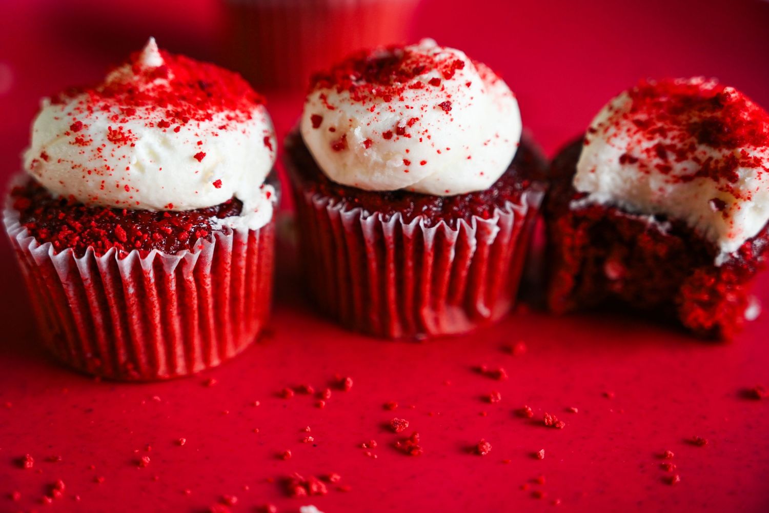 Three red velvet cupcakes with white frosting and red sugar dust on red background.