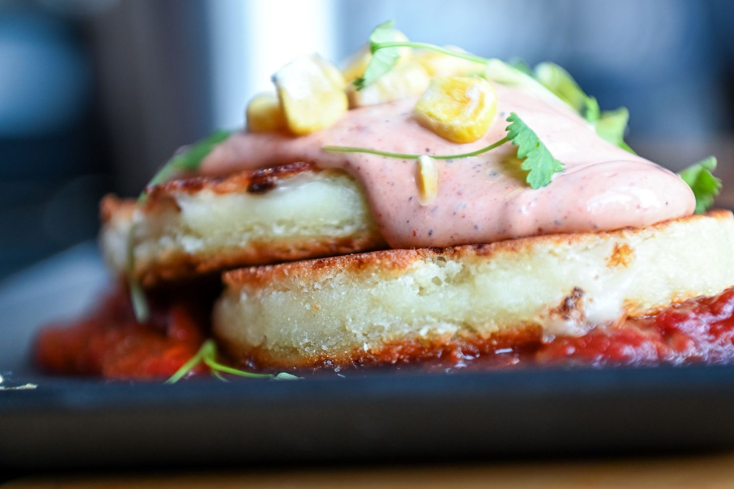 Golden fried sandwich topped with pink sauce, fresh herbs, and citrus on a dark plate.