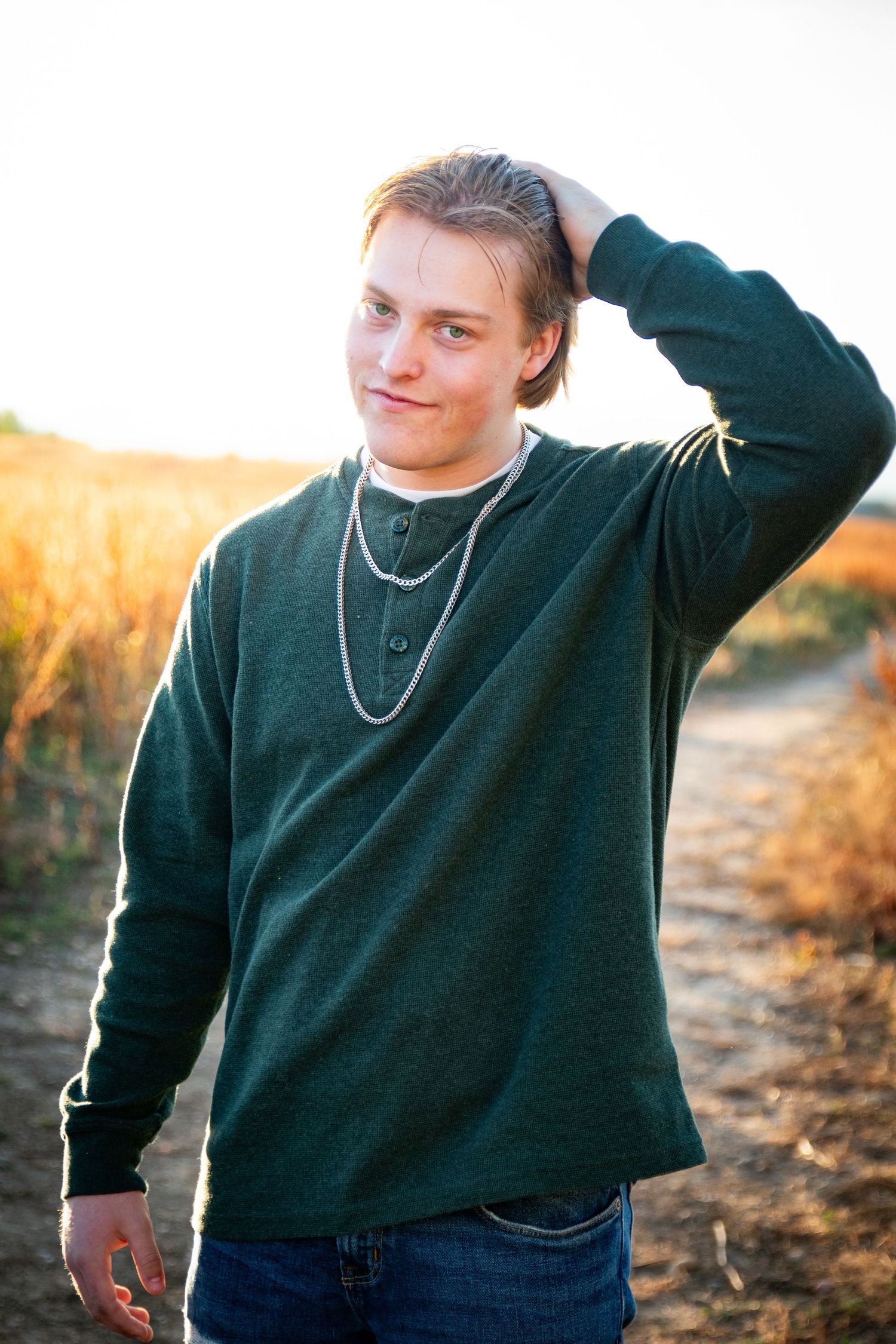 Young man in green henley sweater with chain necklace posing in golden field at sunset.