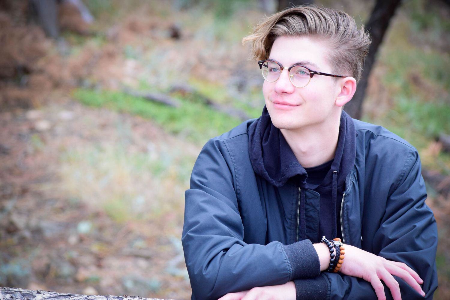 Smiling teenage boy with glasses resting on a log in an autumn outdoor setting.
