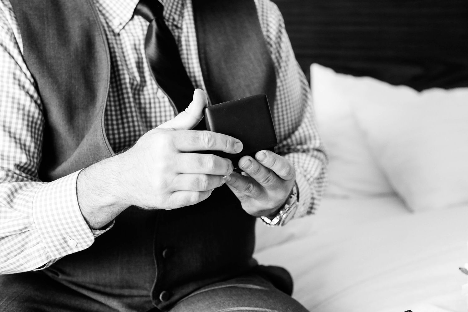 Black and white photo of a man in a vest and tie playing acoustic guitar while seated on a bed.