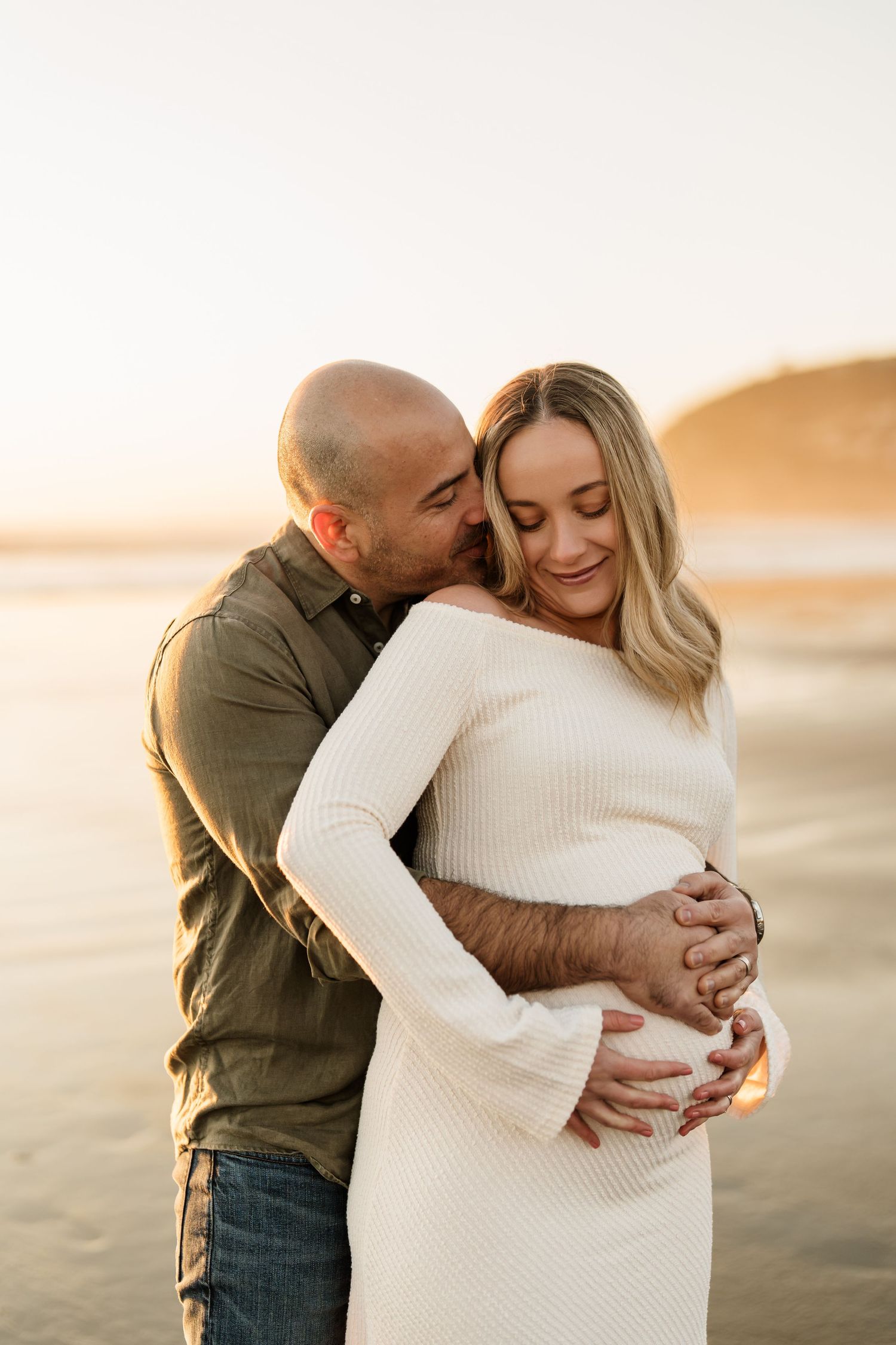Expectant couple embracing in golden sunrise light during their Sumner Beach maternity photoshoot.