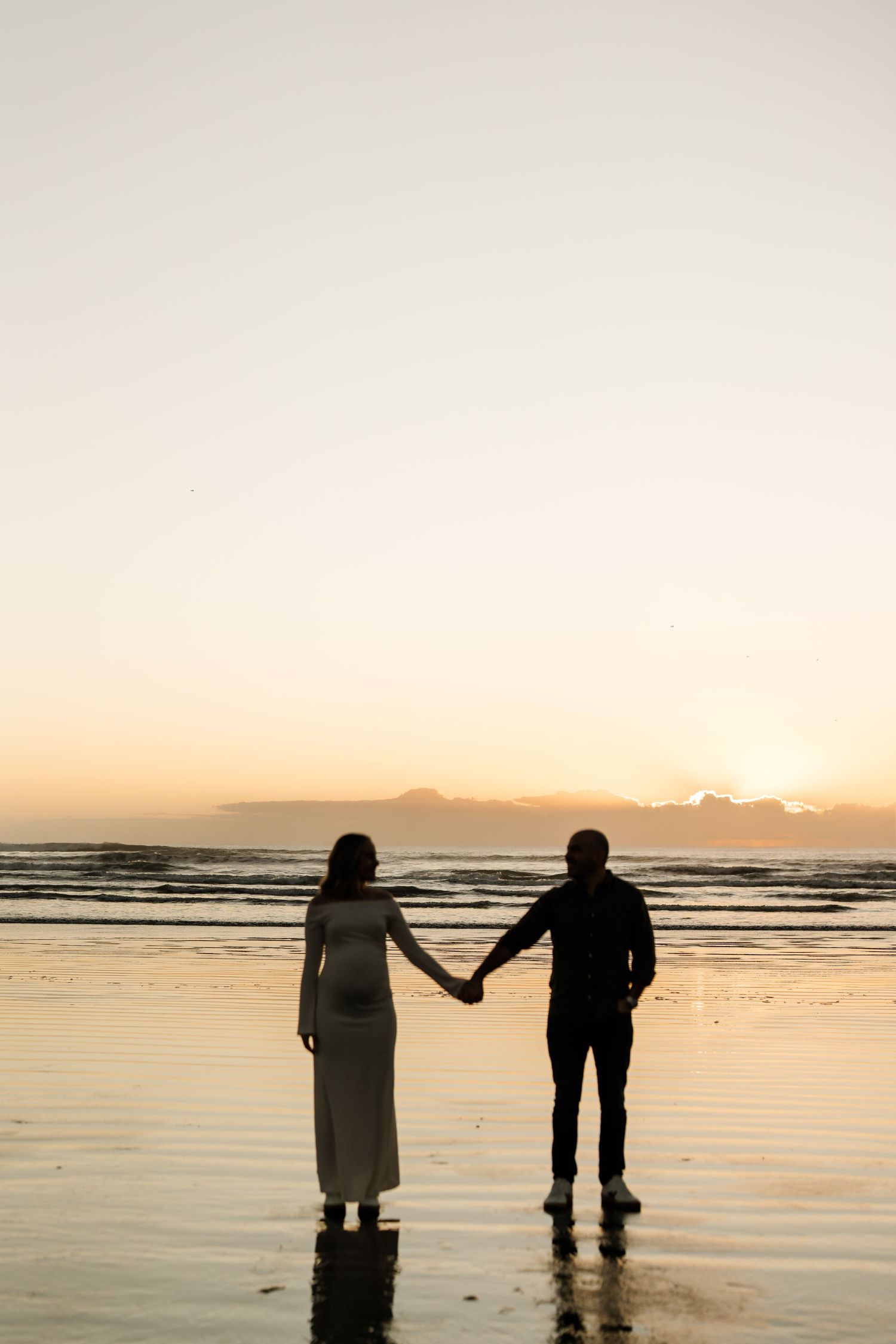 Expectant couple walking hand in hand along Sumner Beach at sunrise during their Christchurch maternity session.