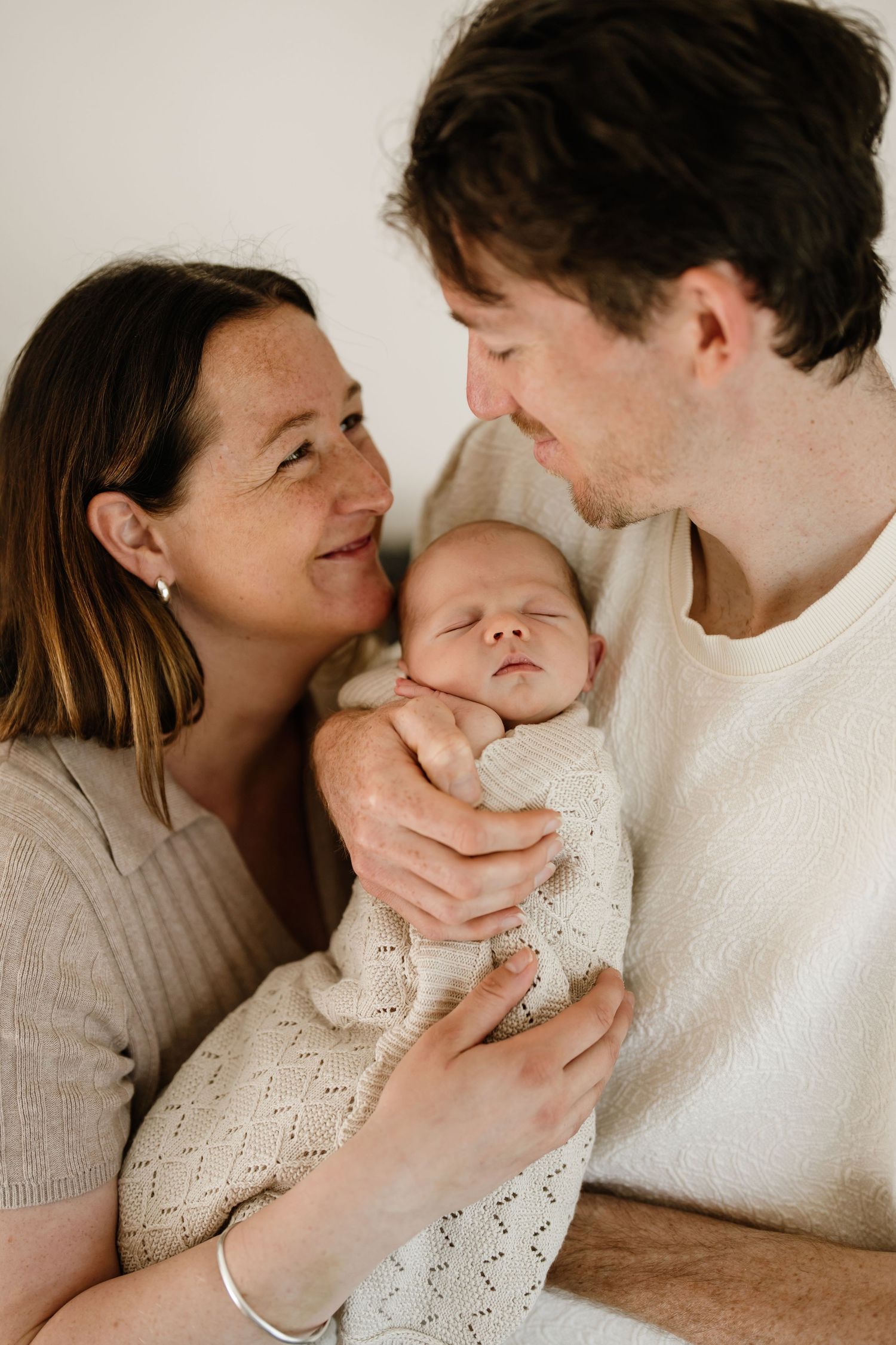 A couple smiling at each other while holding their newborn baby at an in home newborn photography session