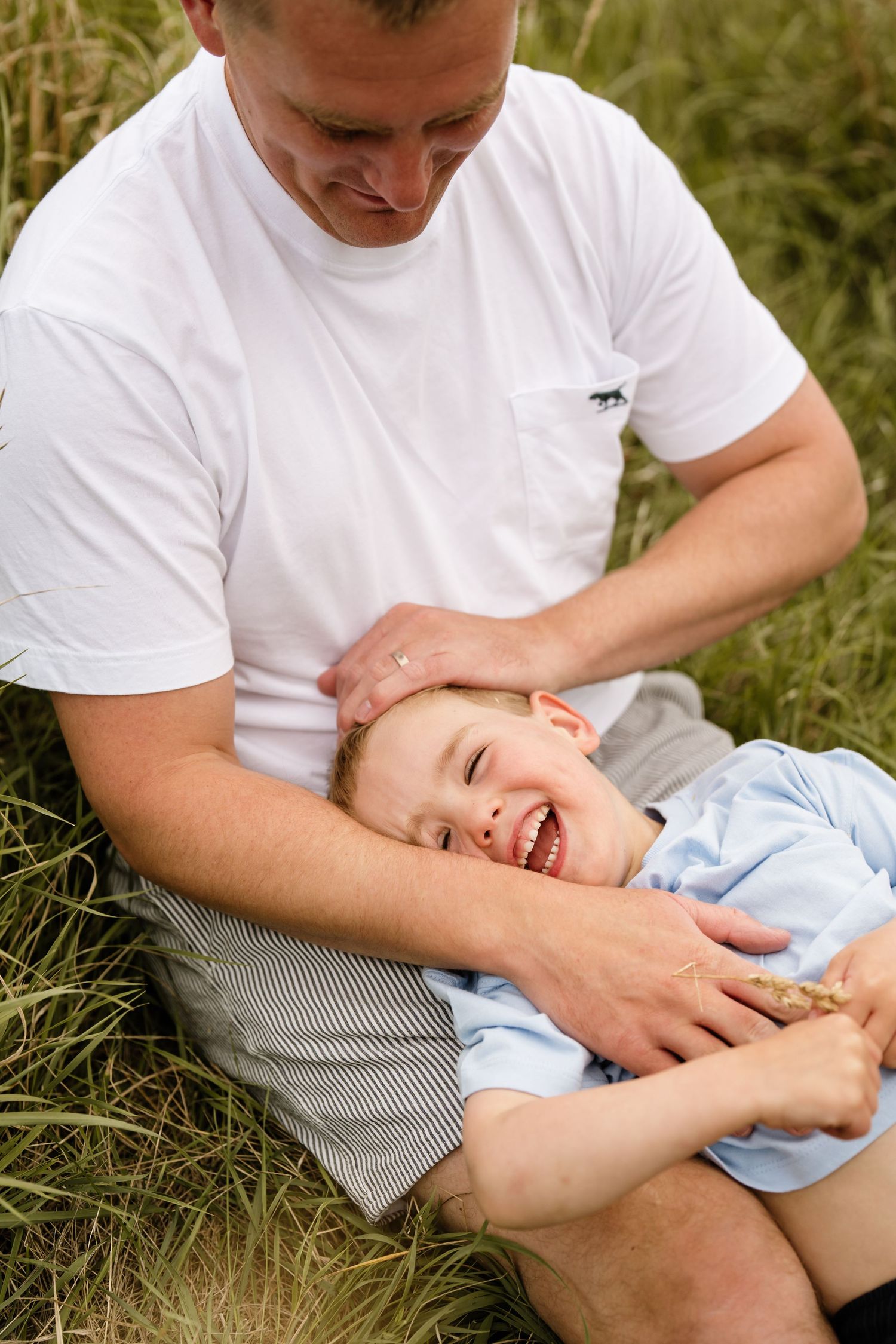 Dad cuddling and playing with his young son in long grass during an outdoor family photography session