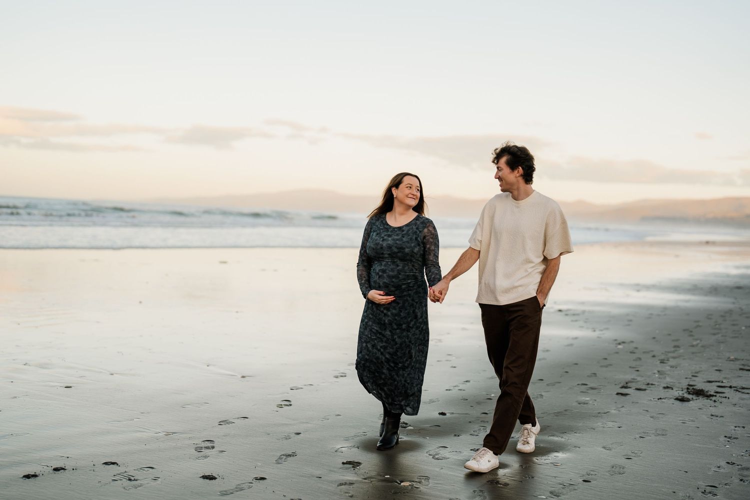A couple smiling at each other on a Christchurch beach at sunset, during a maternity photography session
