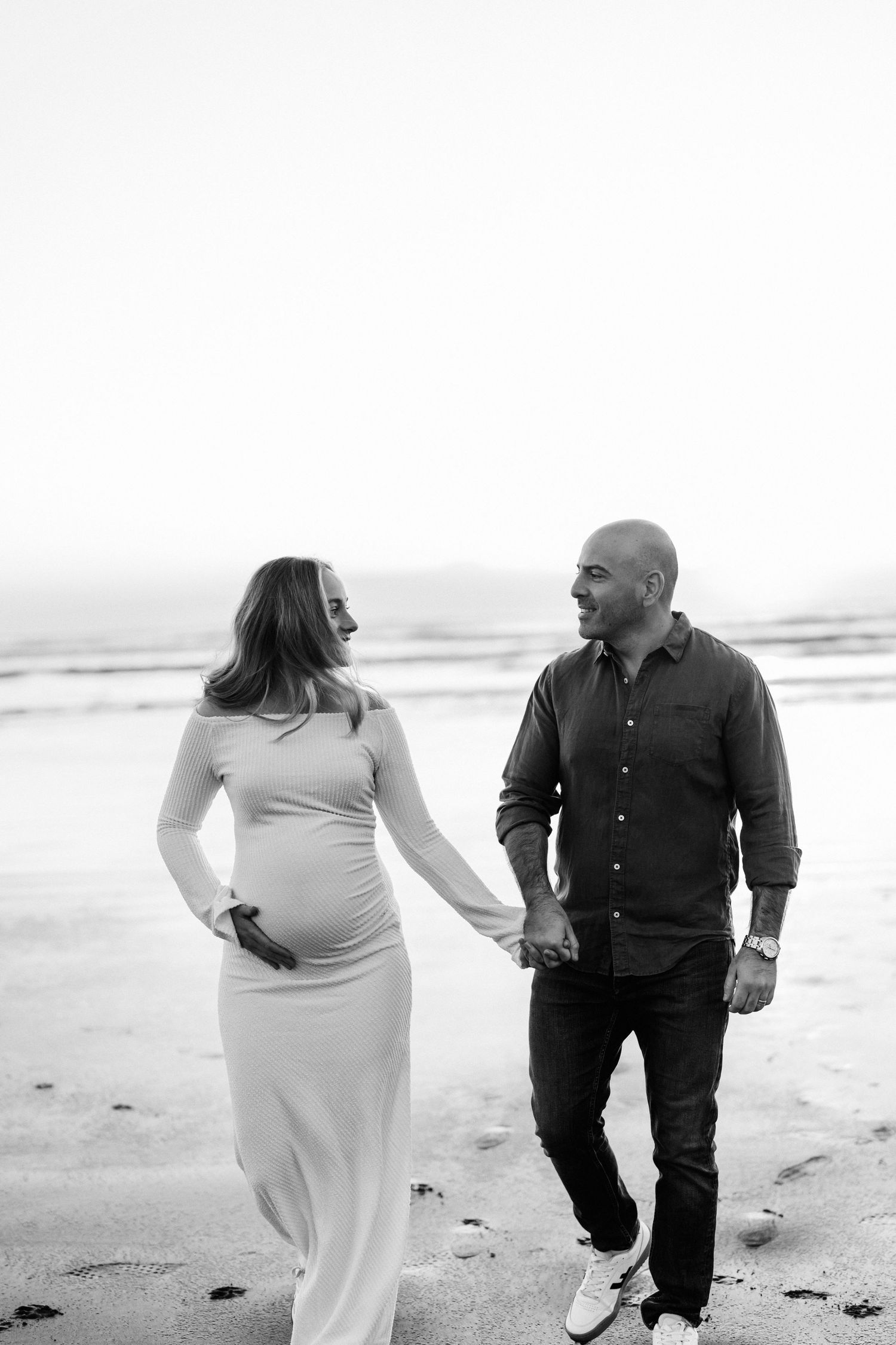 Black and white photo of an expectant couple walking hand in hand along Sumner Beach during their maternity session.