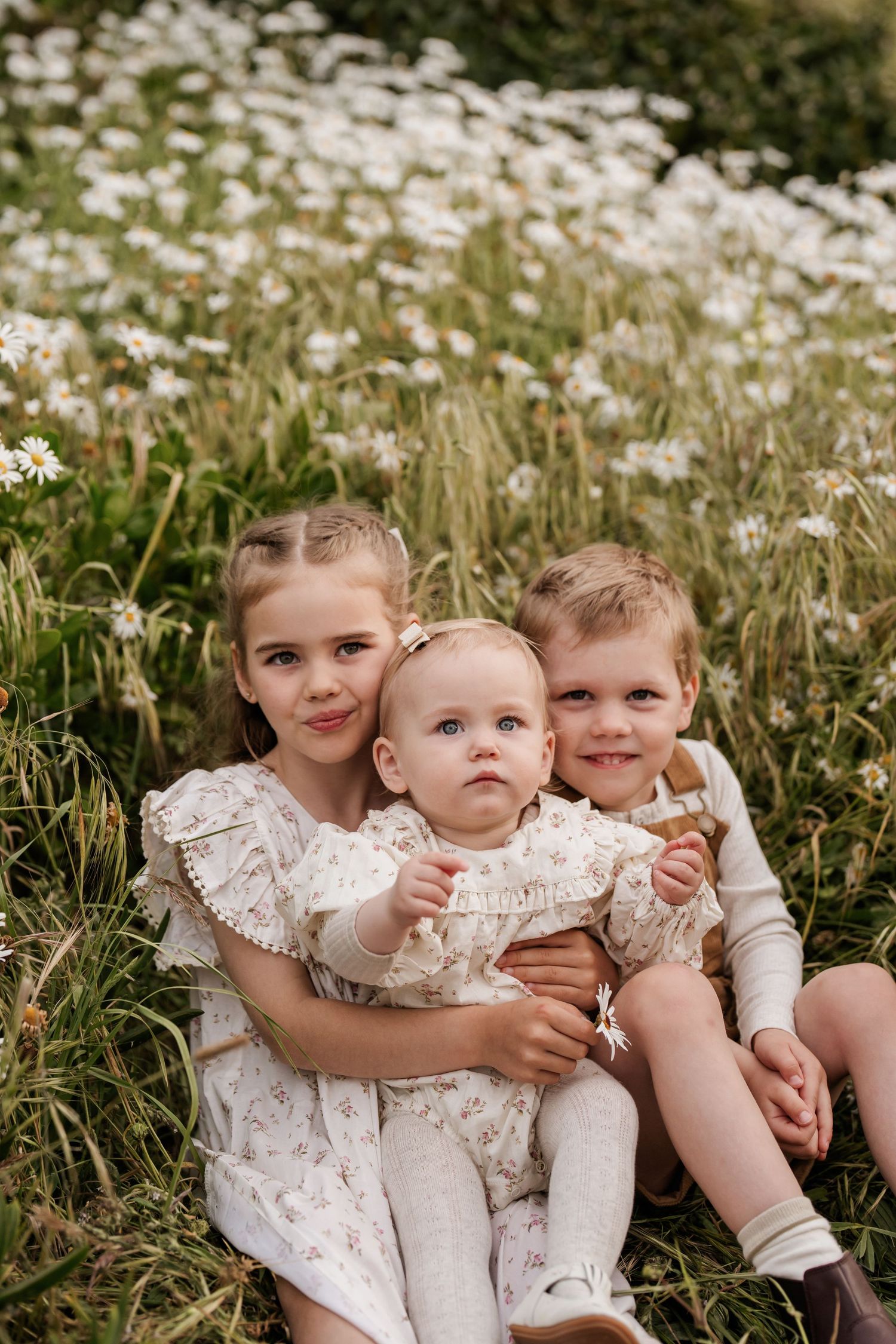 Three young siblings sitting together among wildflowers during an outdoor family photography session in Christchurch
