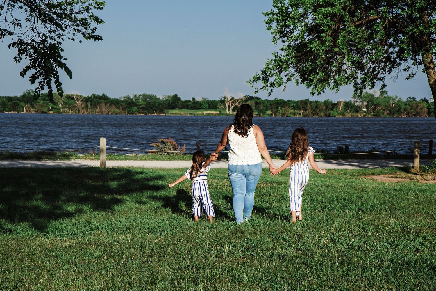 A mother holds hands with two young girls walking toward a scenic lakeside park on a sunny day.