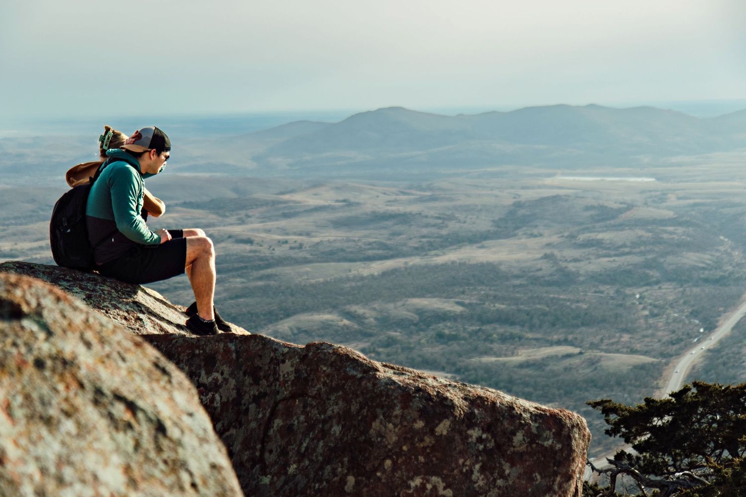 Hiker sits on rocky mountain peak overlooking vast desert landscape with distant mountains in background.