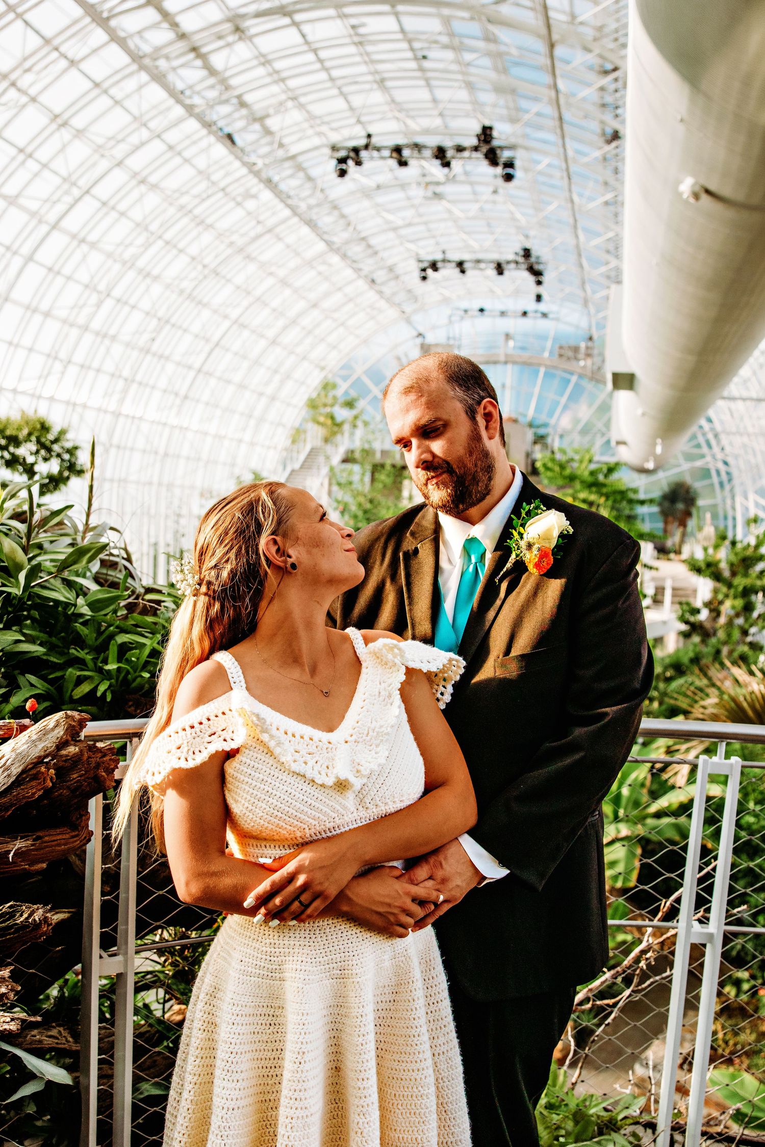 A romantic wedding couple shares intimate moments in a greenhouse conservatory with glass dome ceiling and lush plants.