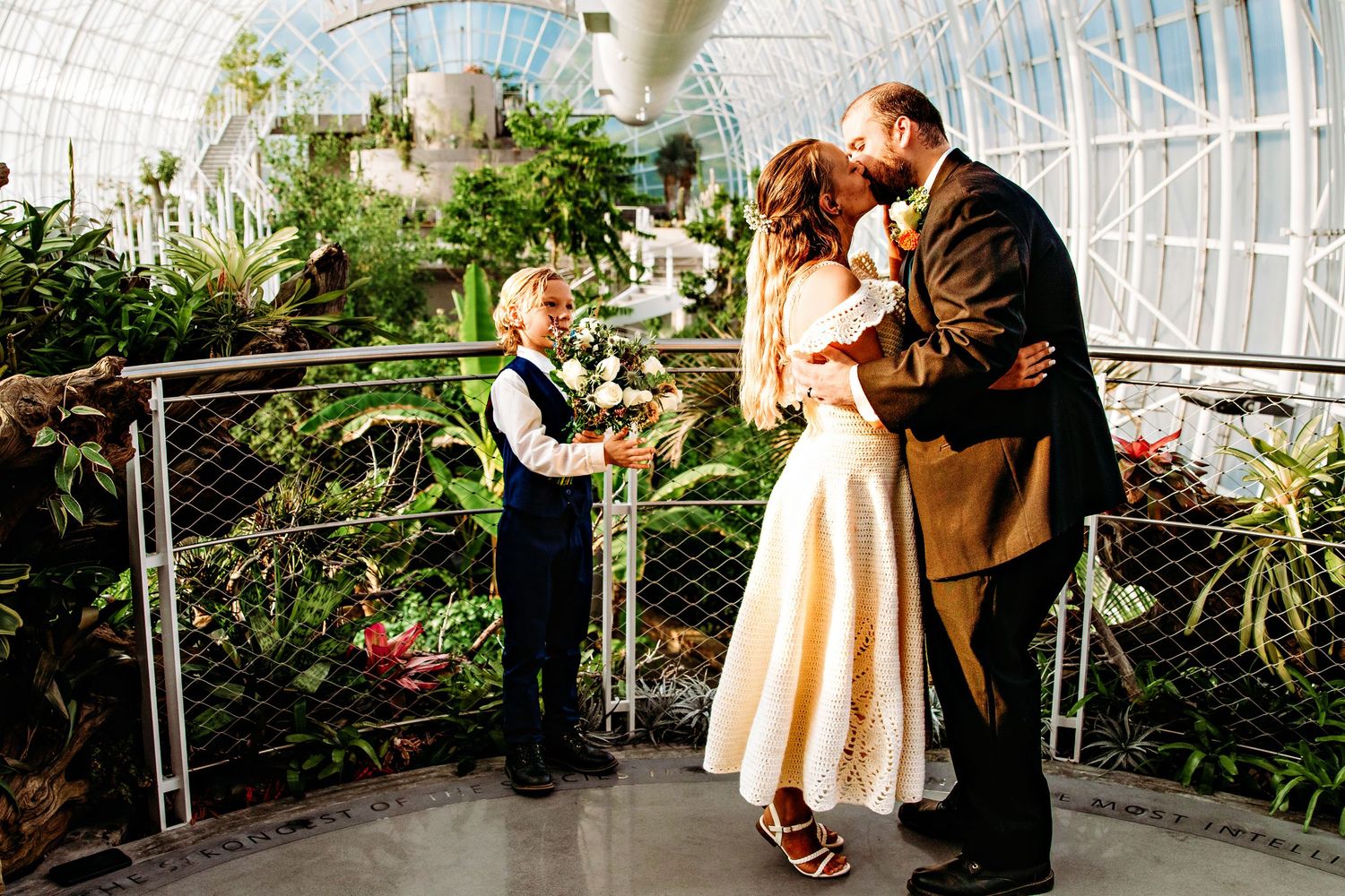 A couple in formal attire poses together in a greenhouse surrounded by lush plants and flowers.