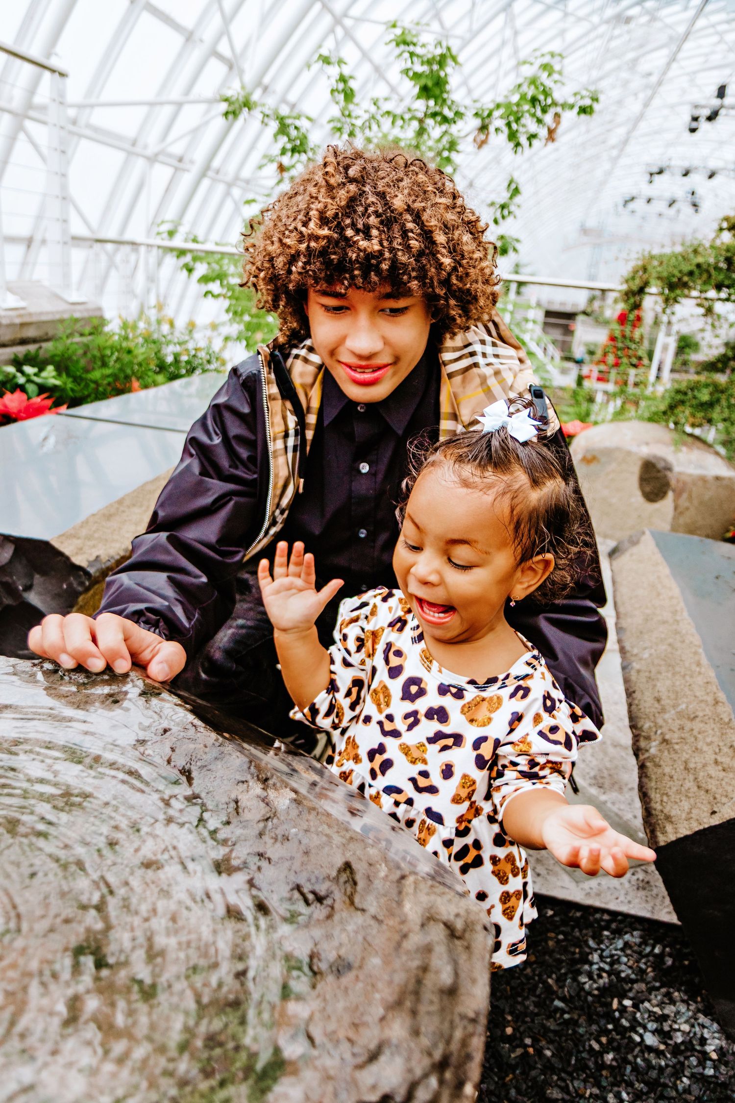 A teen with curly hair and a young girl in leopard print explore a water feature inside a greenhouse.