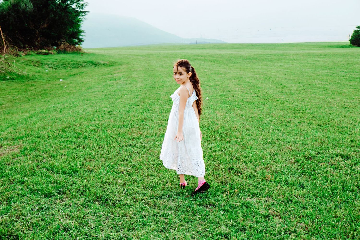 A young girl in a white dress stands in a lush green field with misty mountains in the background.
