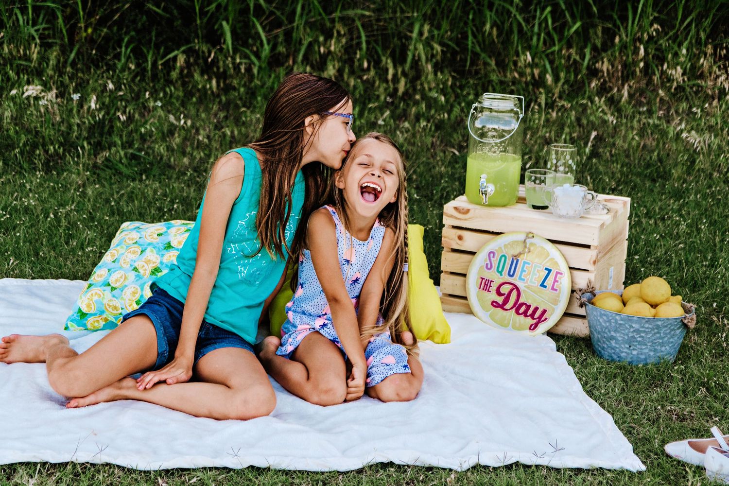 Two girls laughing on a picnic blanket with lemonade stand setup outdoors on a sunny day.