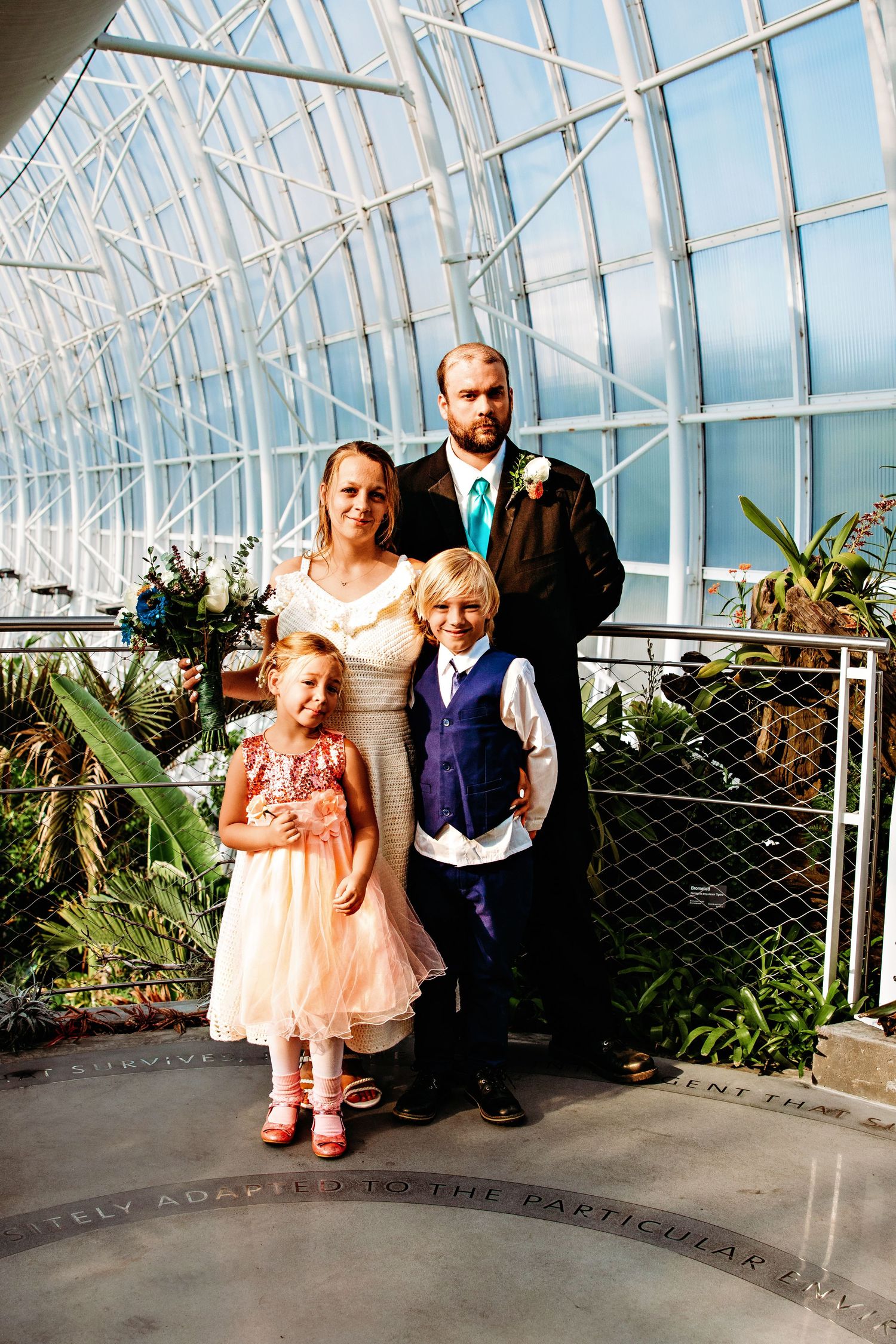 Family poses together in formal attire inside a glass conservatory with tropical plants and natural lighting.