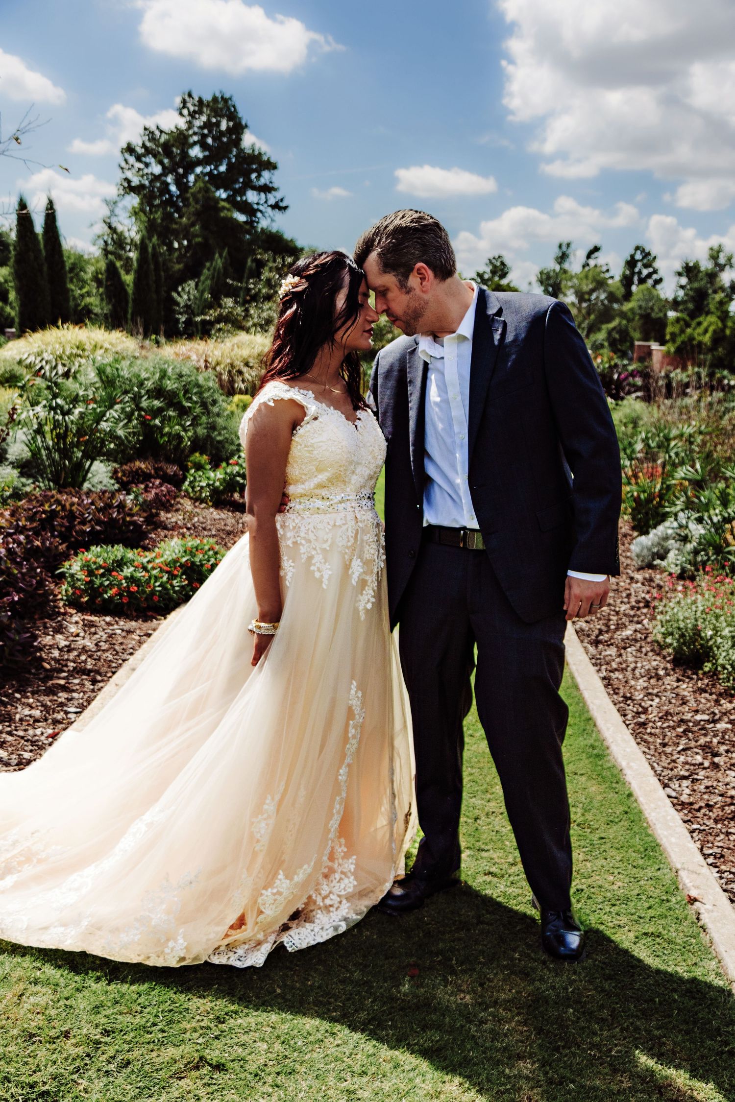 Bride in lace gown and groom in navy suit share romantic moment on garden path on their wedding day.