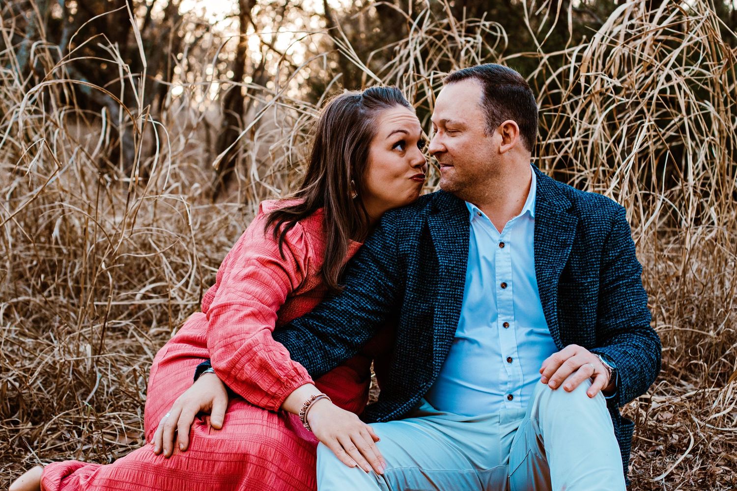 A couple in pink and blue outfits share intimate moments in a field of tall dried grass at sunset.