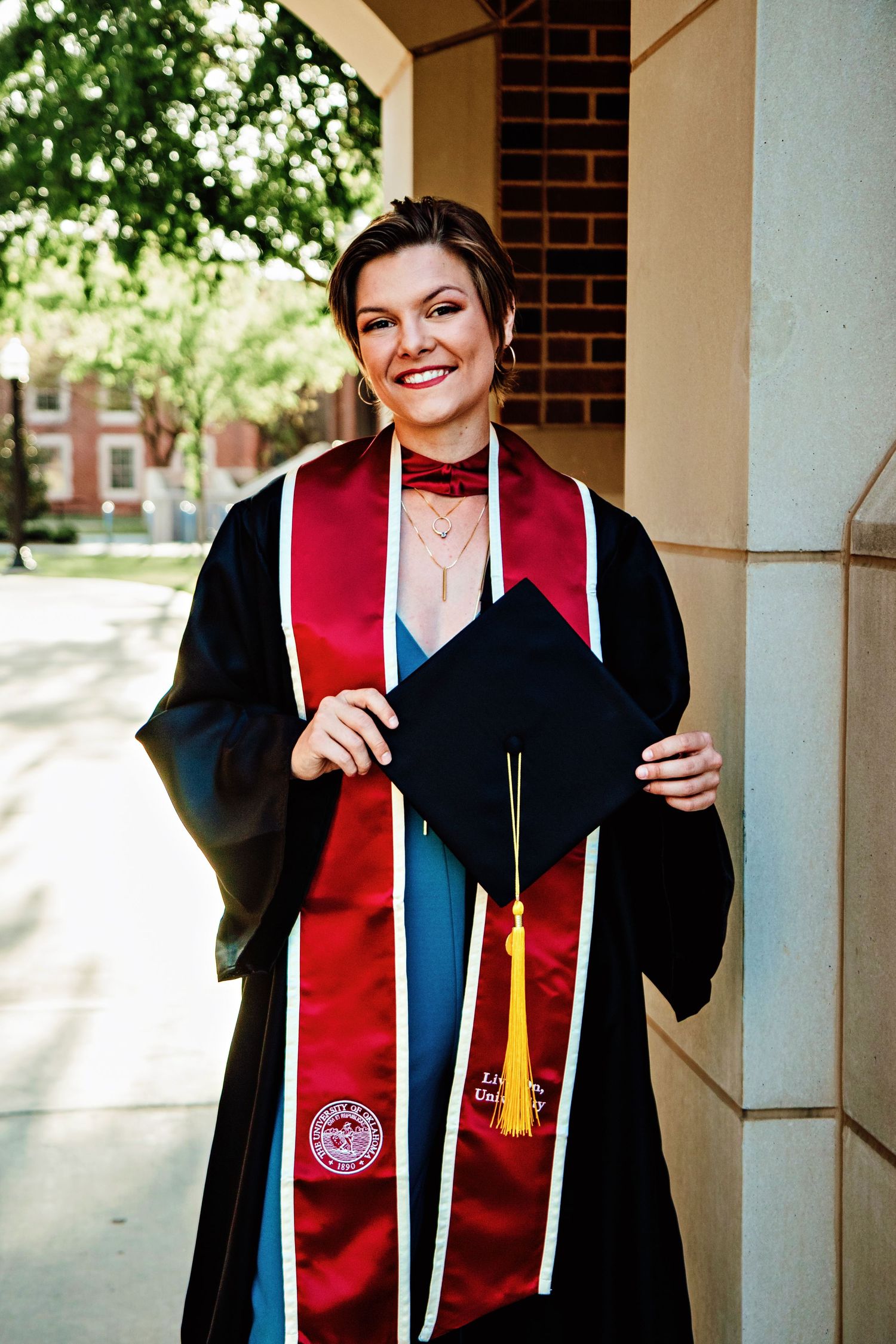 Graduate wearing black and red graduation regalia poses for photos in a brick columned walkway.